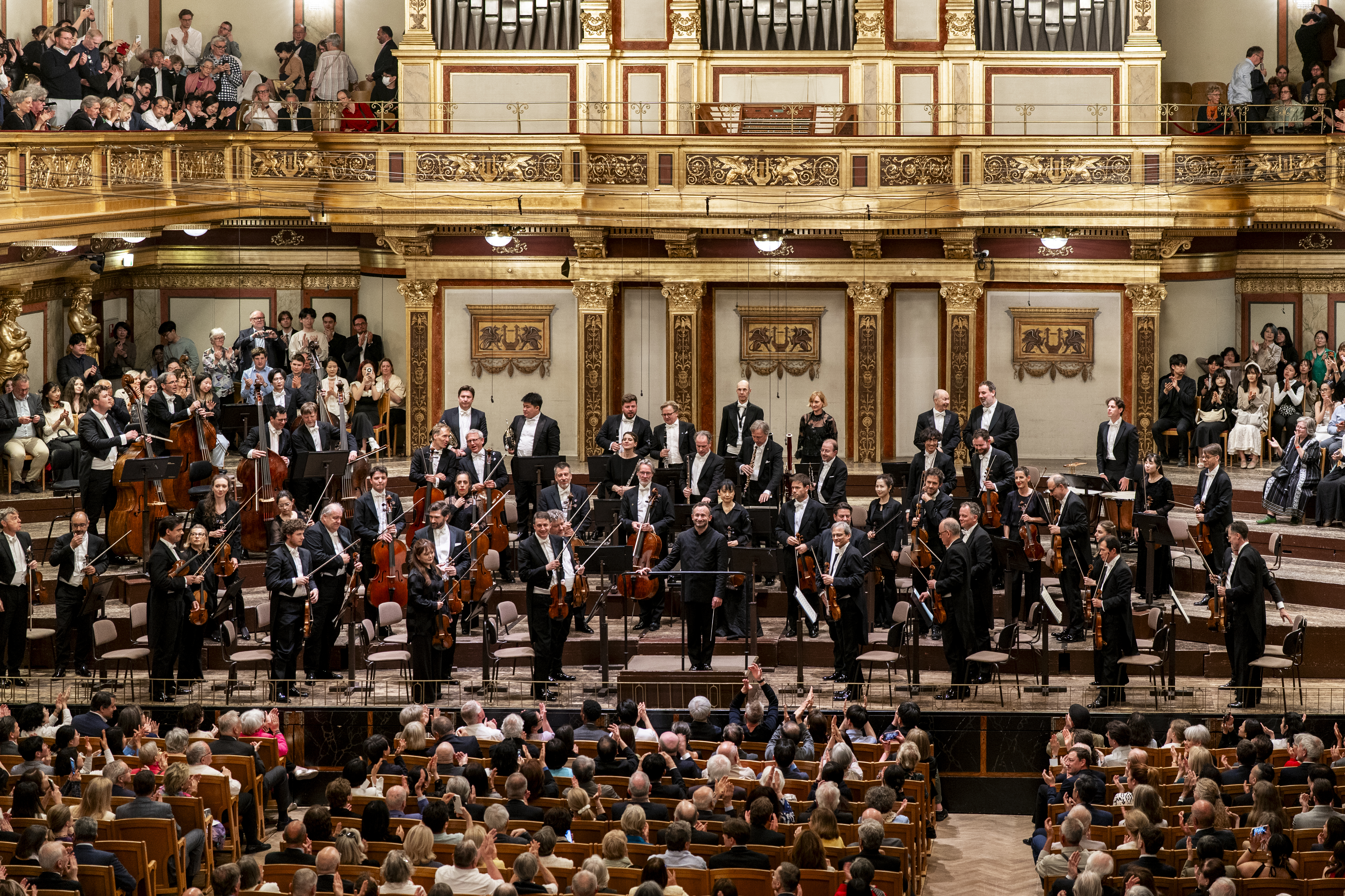 Frontalansicht der gesamten Bühne des Goldenen Saals. Das Orchester hat sich zum Applaus erhoben, während der Dirigent in der Mitte steht. Die beeindruckende goldene Orgel bildet den zentralen Hintergrund dieser symmetrischen Komposition.
