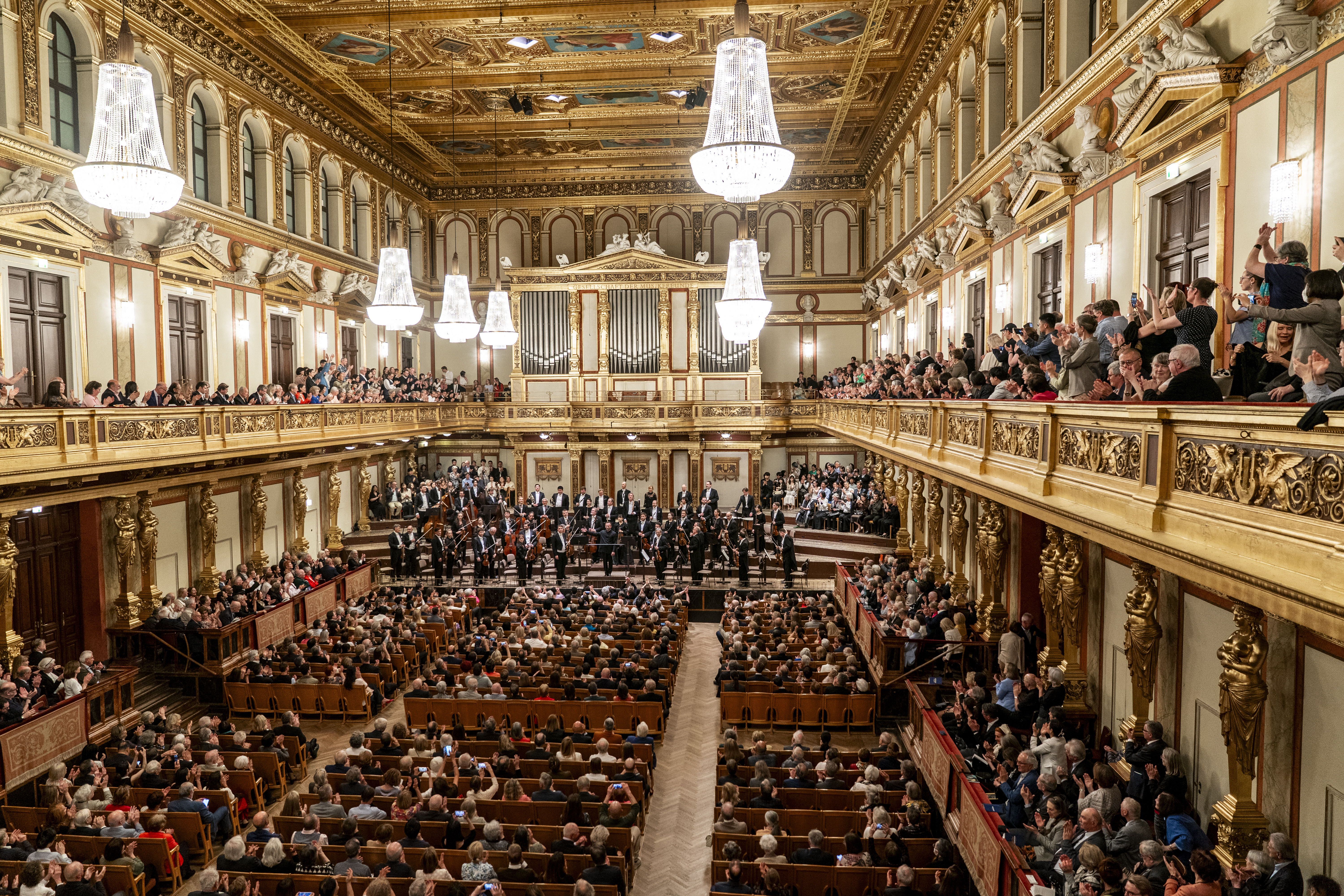 Eine imposante Totalaufnahme des gesamten Goldenen Saals im Wiener Musikverein von einem oberen Rang aus. Der Saal ist voll besetzt, die prunkvolle Kassettendecke mit Gemälden, die riesigen Kristalllüster und die goldene Architektur erzeugen eine majestätische Atmosphäre während des Konzerts.