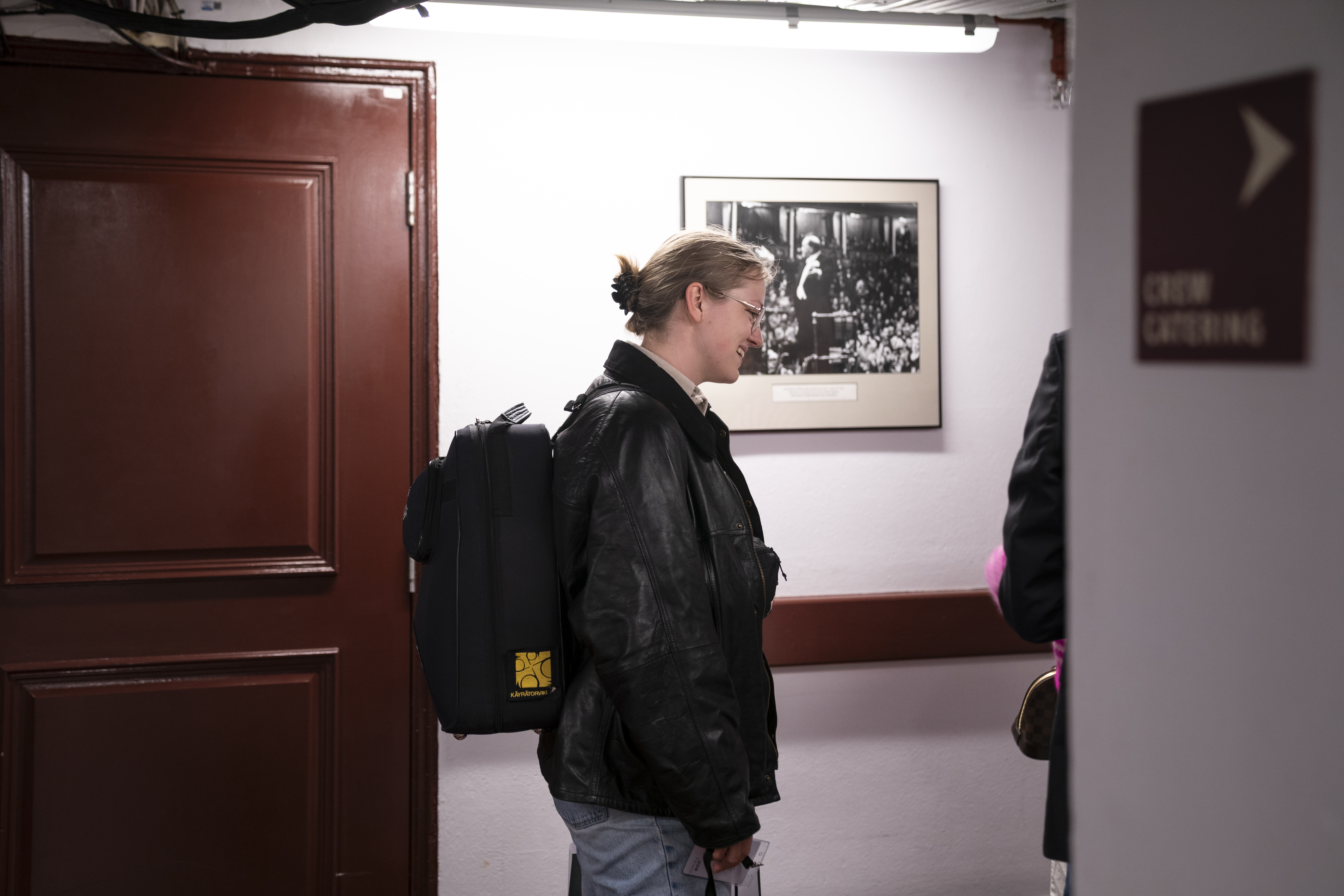 Person with light hair in a ponytail, glasses, a black leather jacket, and a backpack stands near a red door and a framed photo on a wall. A sign reading CREW CATERING is visible.