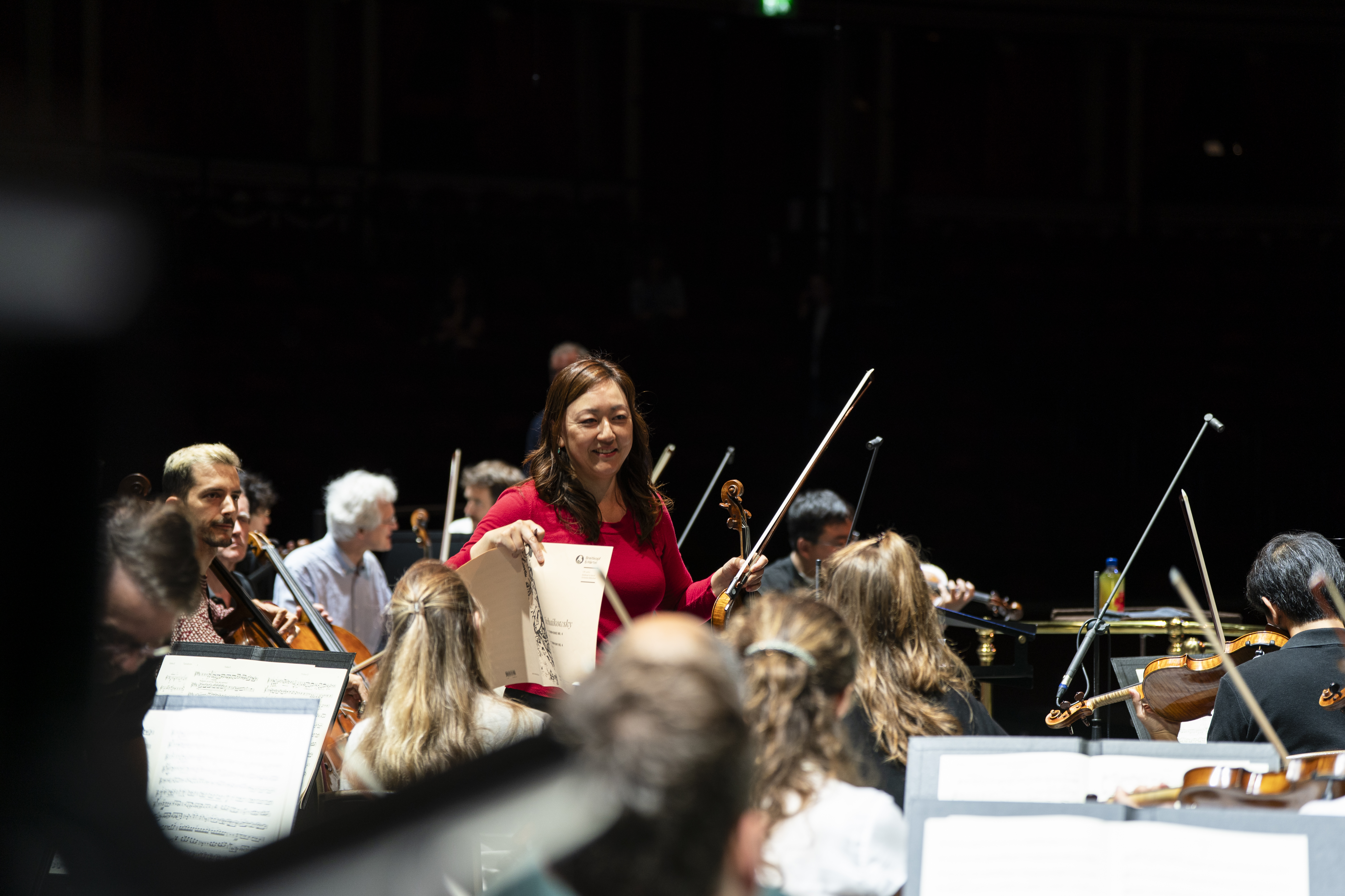 A female violinist in a red top holds her sheet music and violin bow while standing in front of a symphony orchestra on a dark stage during a rehearsal.