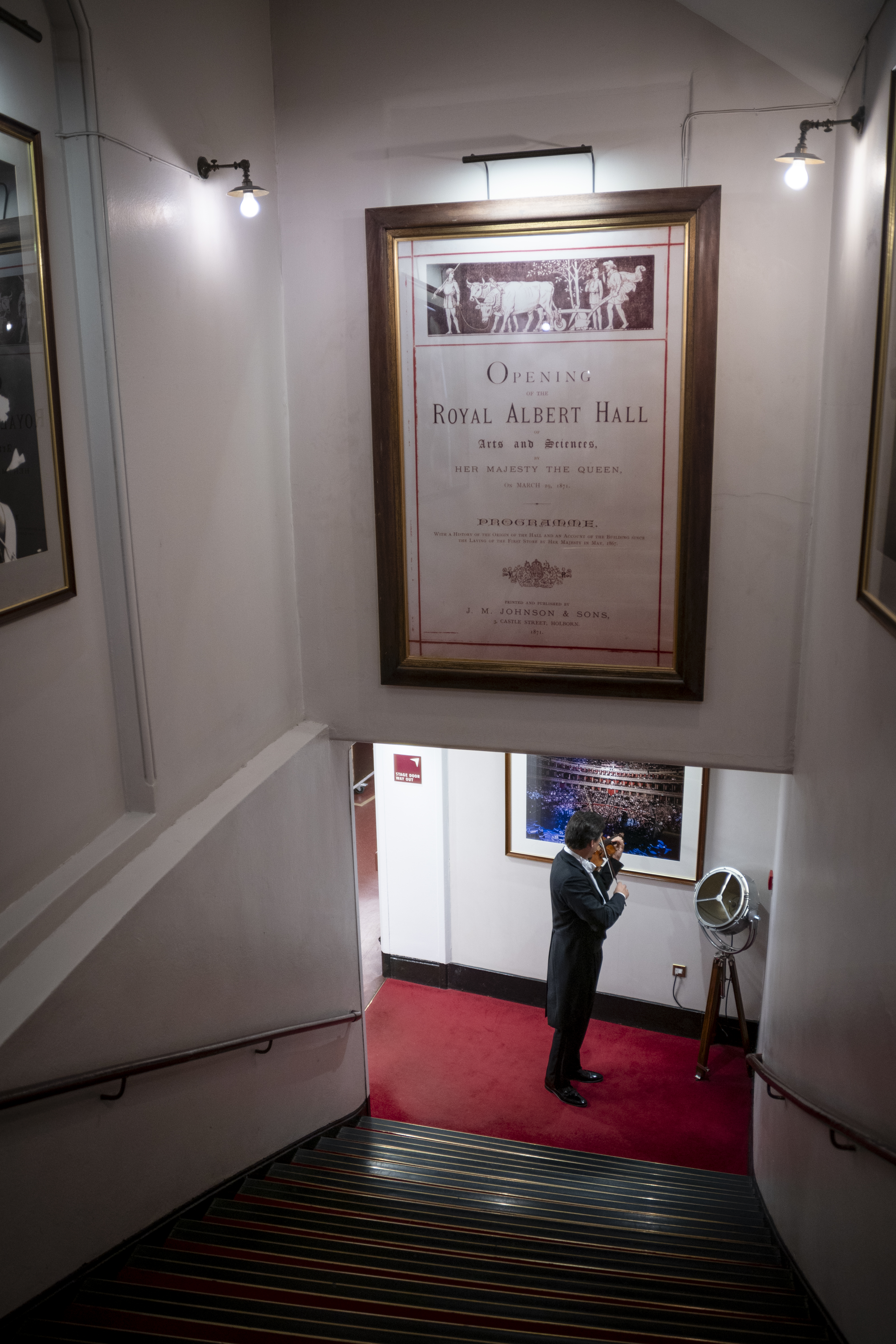 A person in formal attire stands in a hallway beneath a large framed Royal Albert Hall poster, using a payphone next to a red carpeted stairwell.