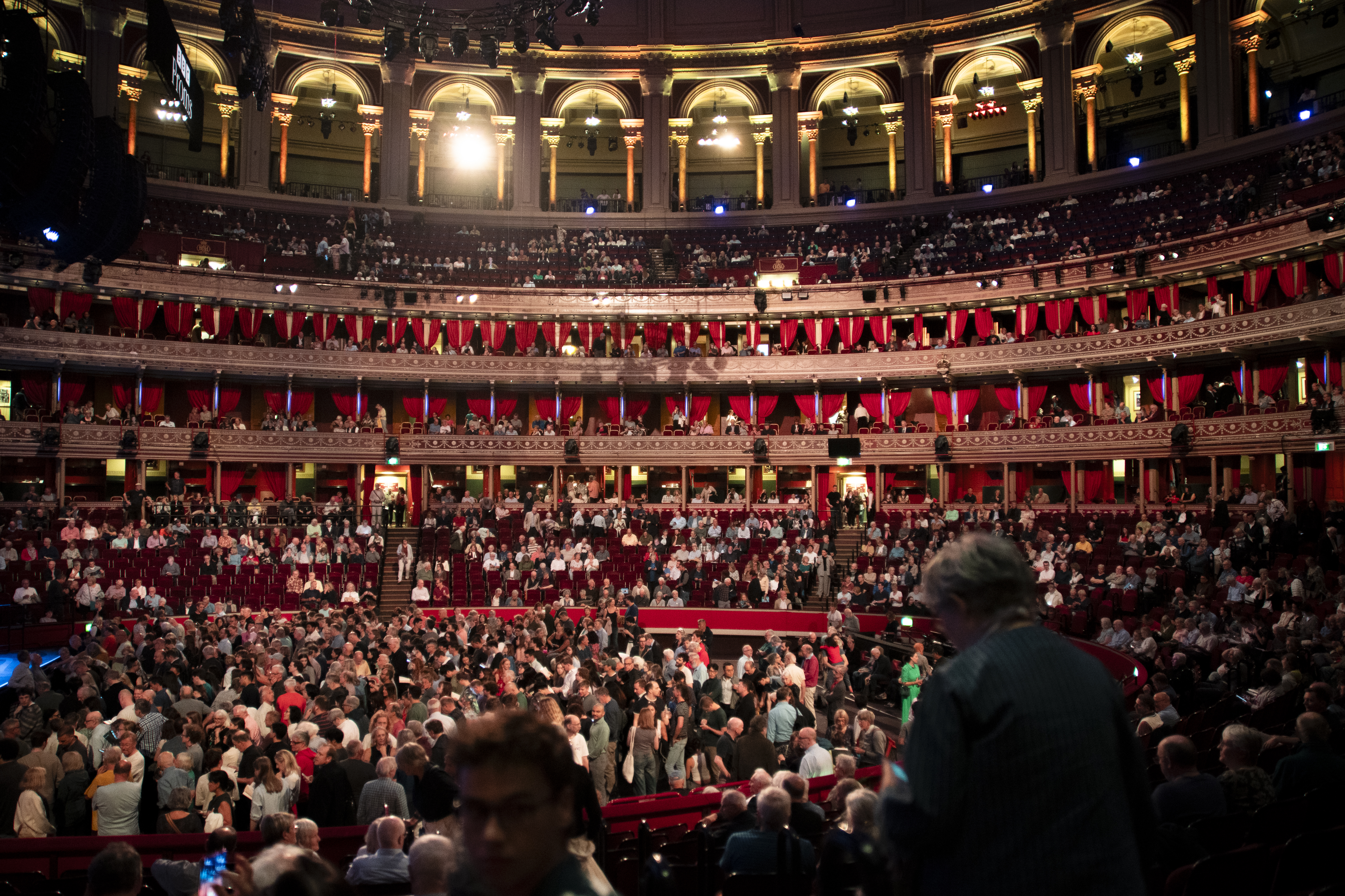 The Royal Albert Hall in London, a grand Victorian venue, is filled with people and hosts the BBC Proms, as shown by the banner in the upper left of the multi-tiered auditorium.