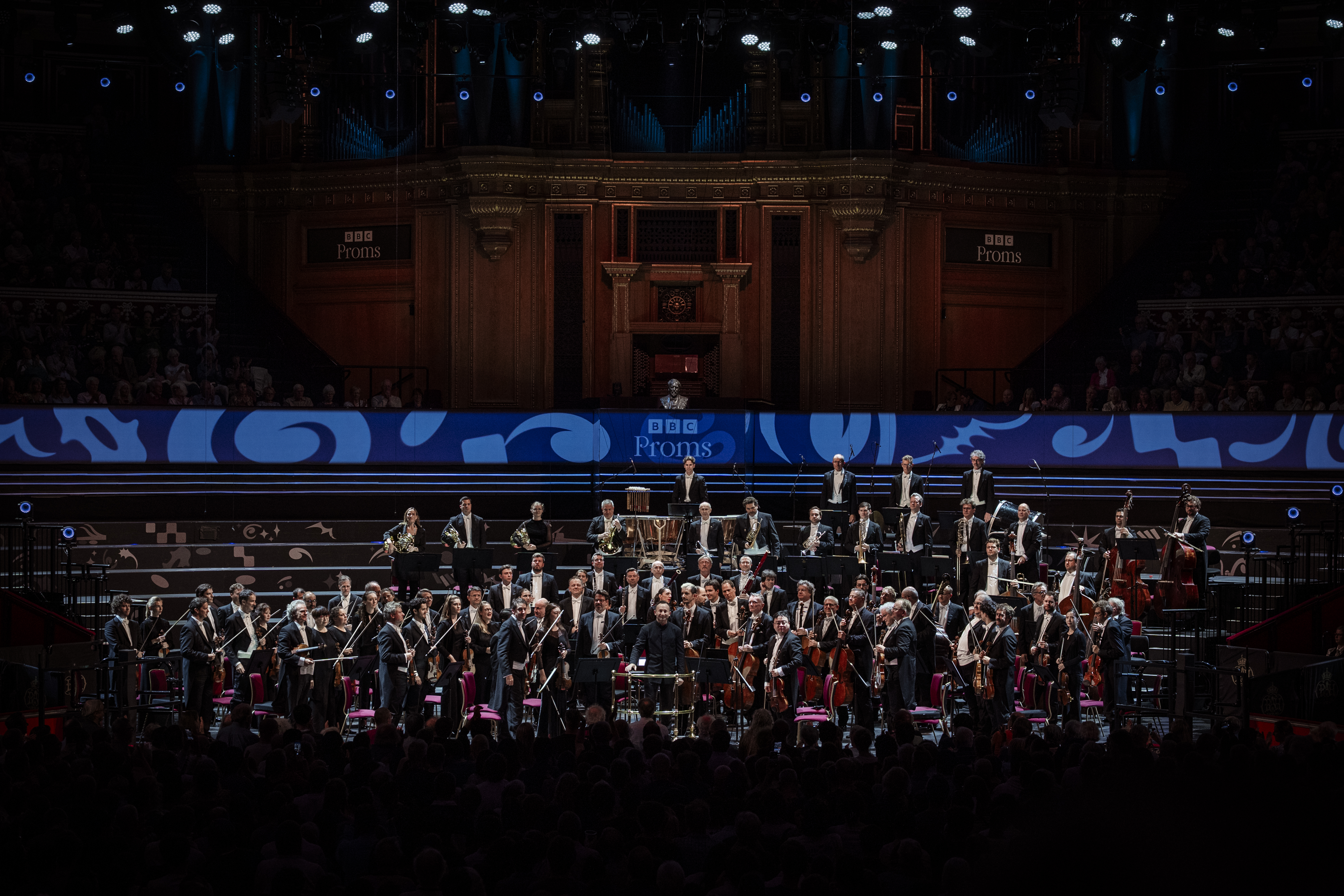 A large symphony orchestra performs on stage at the BBC Proms, with musicians holding various instruments under dramatic lighting in a grand concert hall.