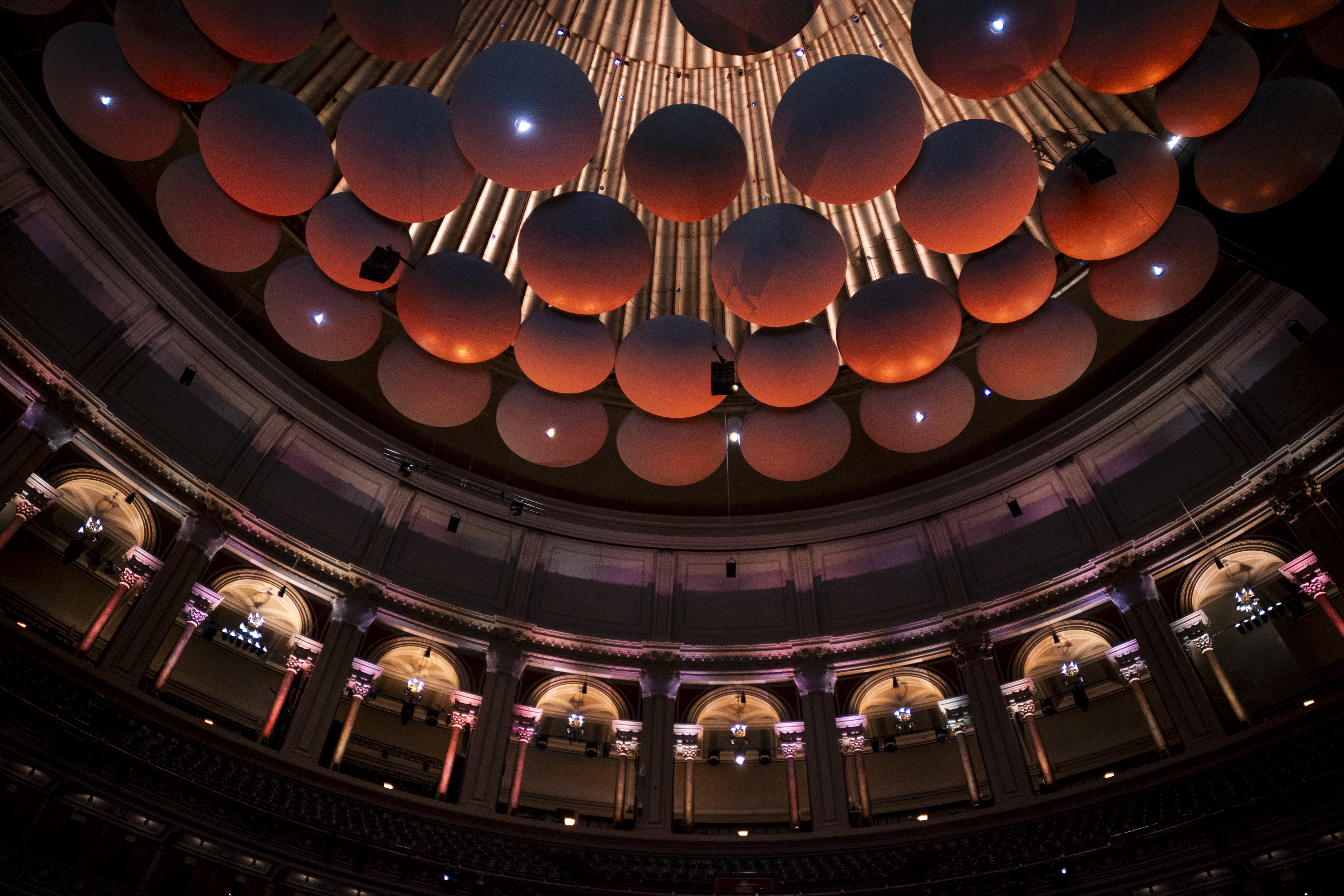 The ornate interior of a concert hall, viewed from below, shows circular acoustic panels and pipe organ pipes on the ceiling, with lit arches and balconies surrounding the space.