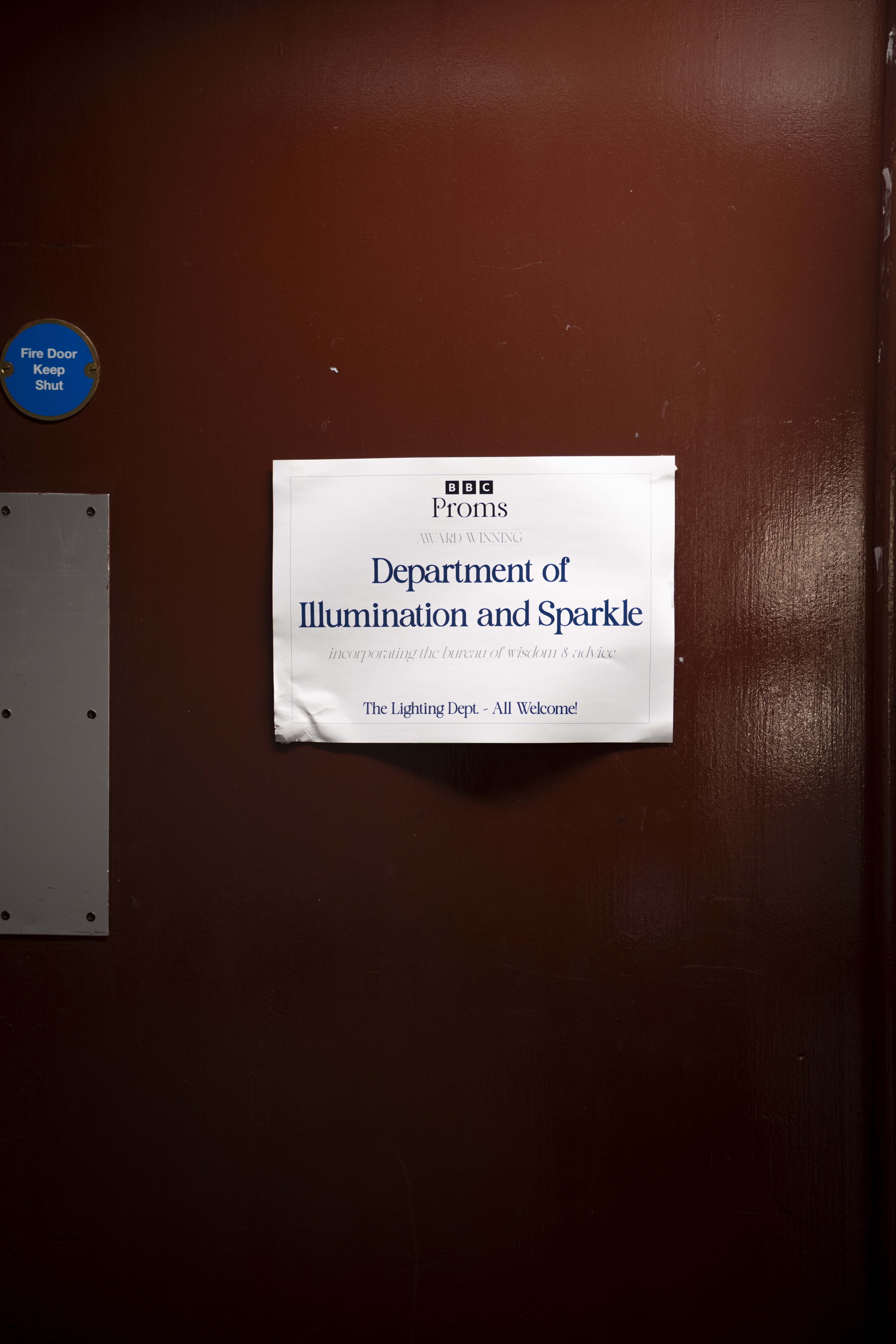 A white sign on a brown door reads: BBC Proms. Department of Illumination and Sparkle. The Lighting Dept. – All Welcome! A blue Fire Door Keep Shut sign is also visible.