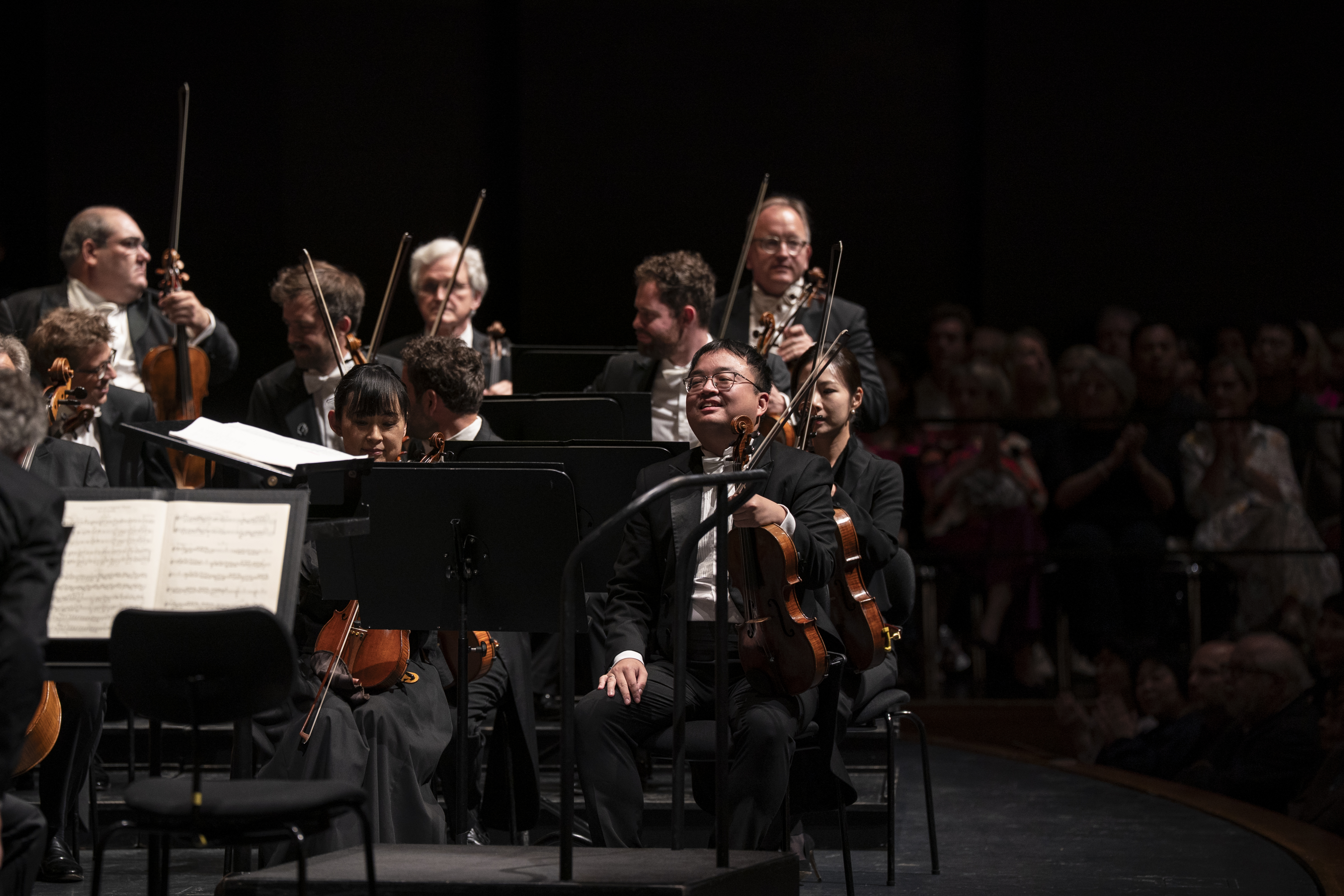 Eine Gruppe von Orchestermusikern in Abendgarderobe sitzt lächelnd mit ihren Instrumenten auf der Bühne, während im Hintergrund das Publikum zusieht.