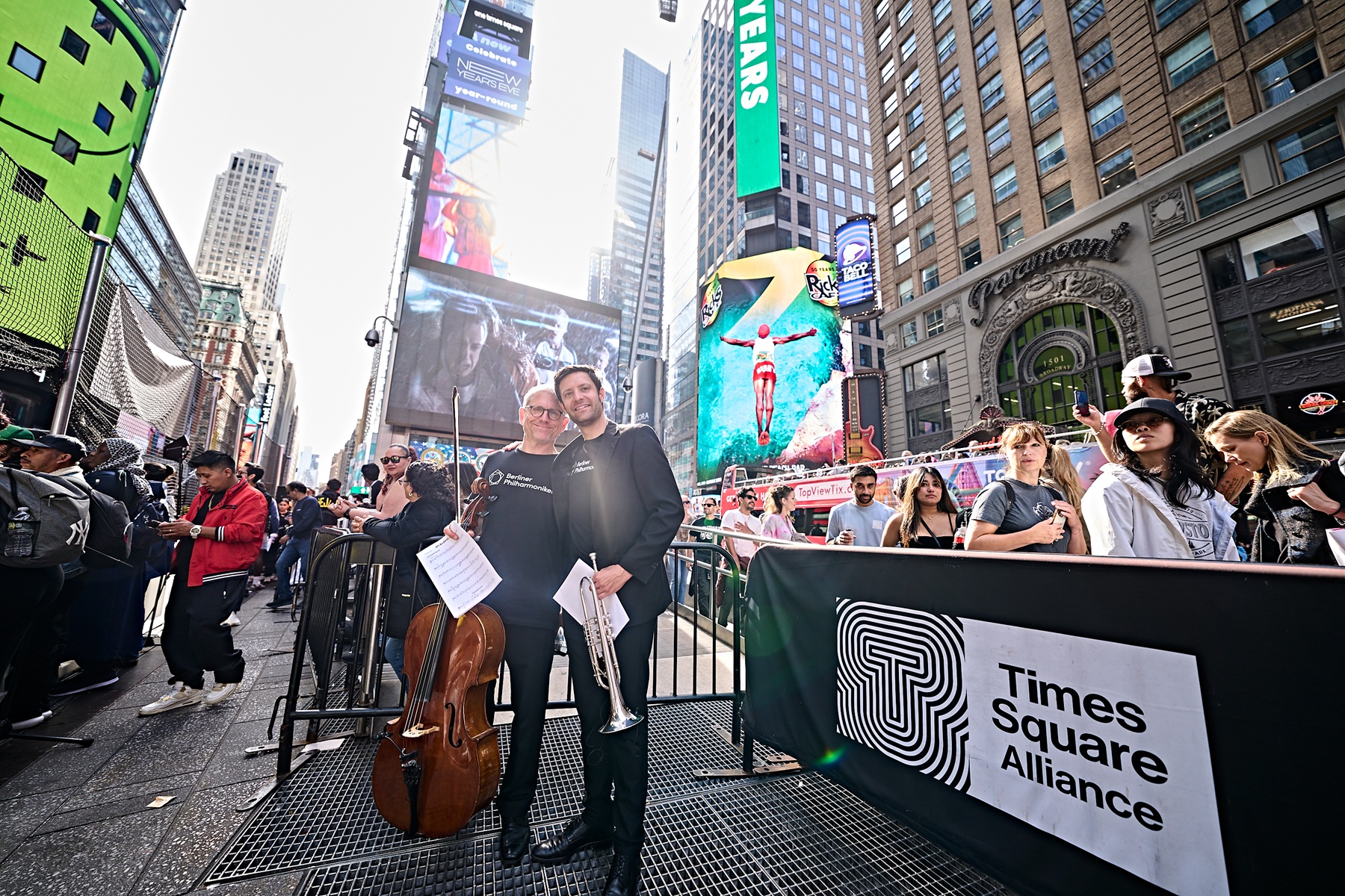 Ein Cellist und ein Trompeter posieren gemeinsam auf einem Bürgersteig am Times Square, umgeben von Menschenmengen und großen digitalen Werbetafeln.