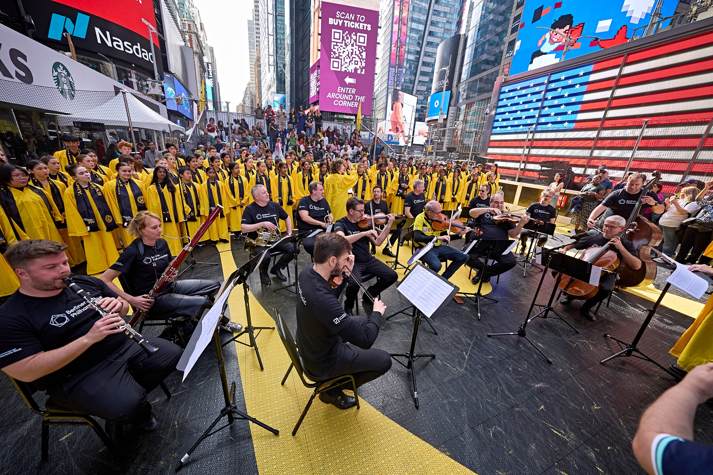 Ein Orchester spielt auf dem Times Square, hinter ihm steht ein großer Chor in gelben Gewändern.
