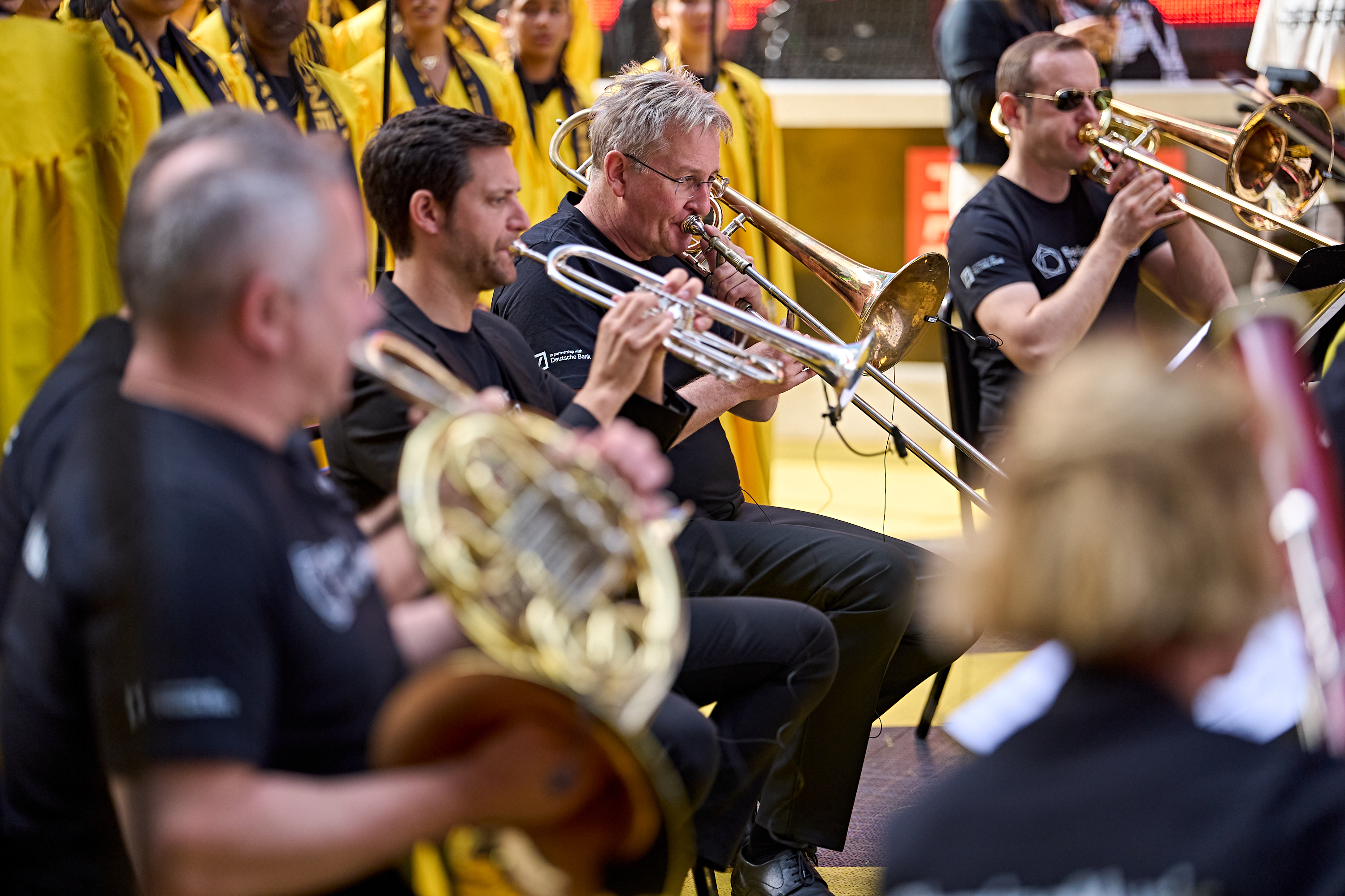 Mitglieder eines Orchesters, die Blechblasinstrumente spielen, darunter eine Trompete und eine Posaune; im Hintergrund sind Chormitglieder in gelben Gewändern zu sehen.
