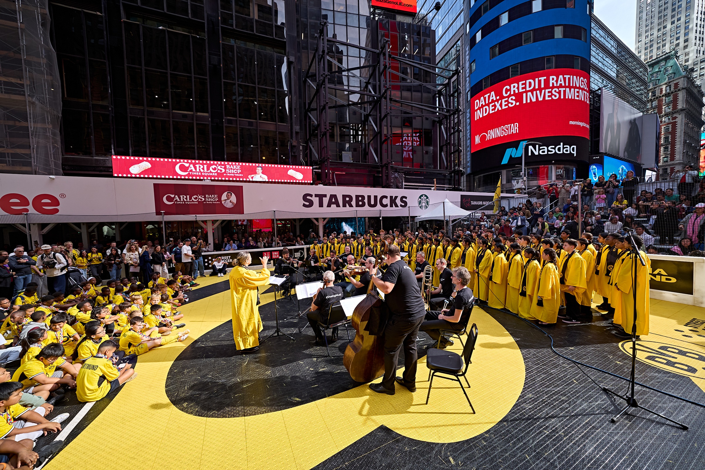 Eine große Gruppe von Studenten in einheitlichen gelben Talaren im Stil von Abschlussroben tritt unter der Leitung eines Dirigenten als Orchester auf einer gelb-schwarzen Bühne am Times Square auf.