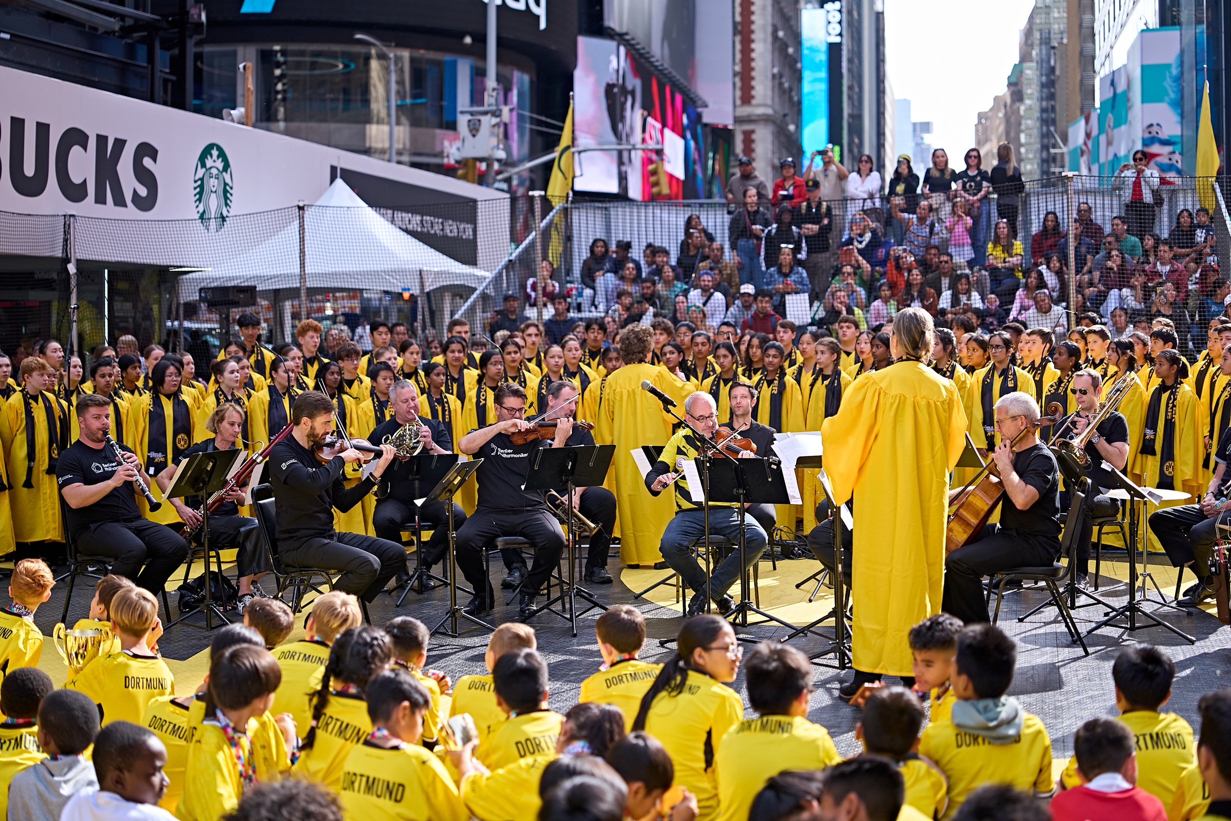 Ein Orchester, dessen Mitglieder größtenteils gelbe T-Shirts tragen, spielt auf dem Times Square vor einem großen Publikum aus Kindern, die ebenfalls gelbe T-Shirts tragen.