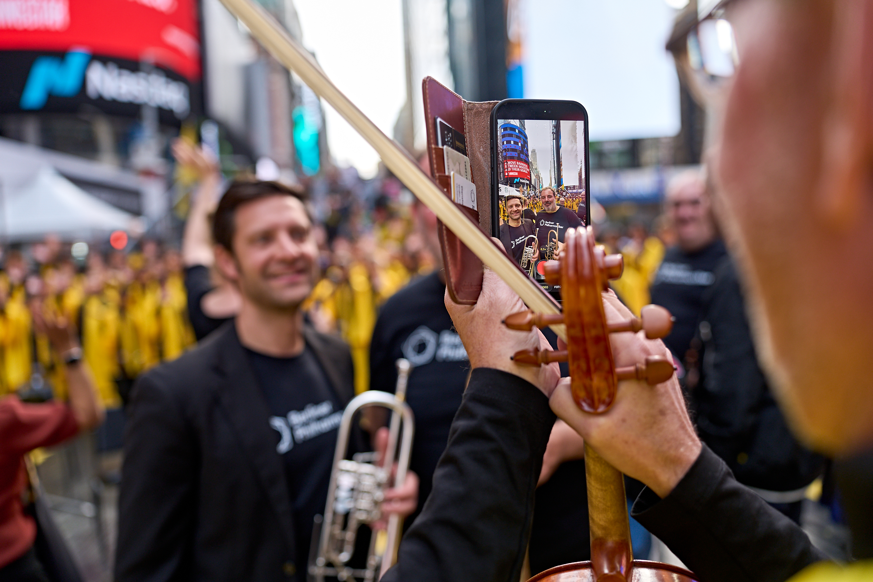 Ein Musiker hält ein Smartphone in der Hand, um ein Foto von zwei anderen Musikern am Times Square zu machen, während er in der anderen Hand einen Geigenbogen hält.