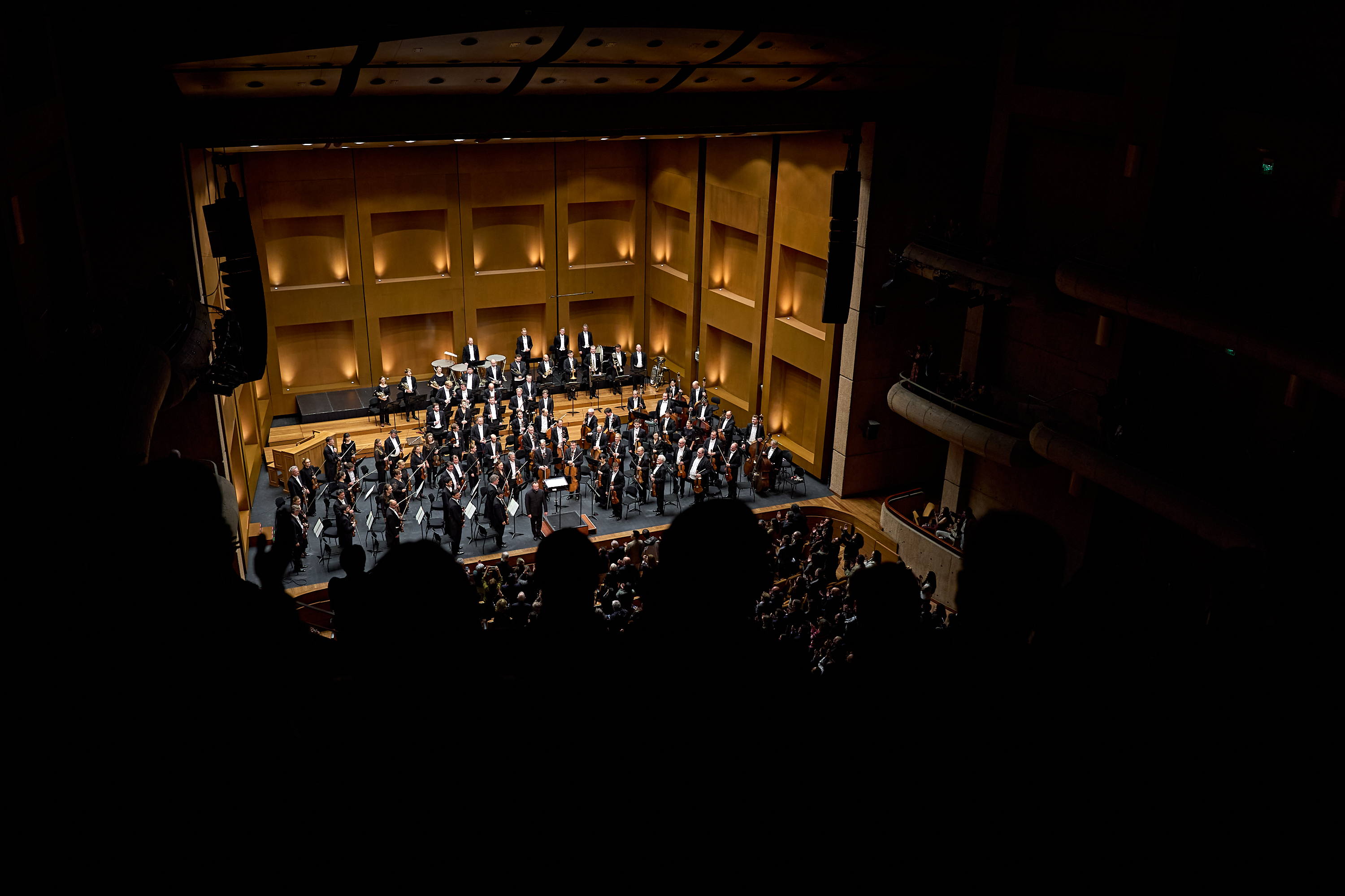 Ein vollbesetztes Orchester spielt auf einer hell beleuchteten Bühne in einem dunklen Konzertsaal, gesehen aus dem Zuschauerraum.