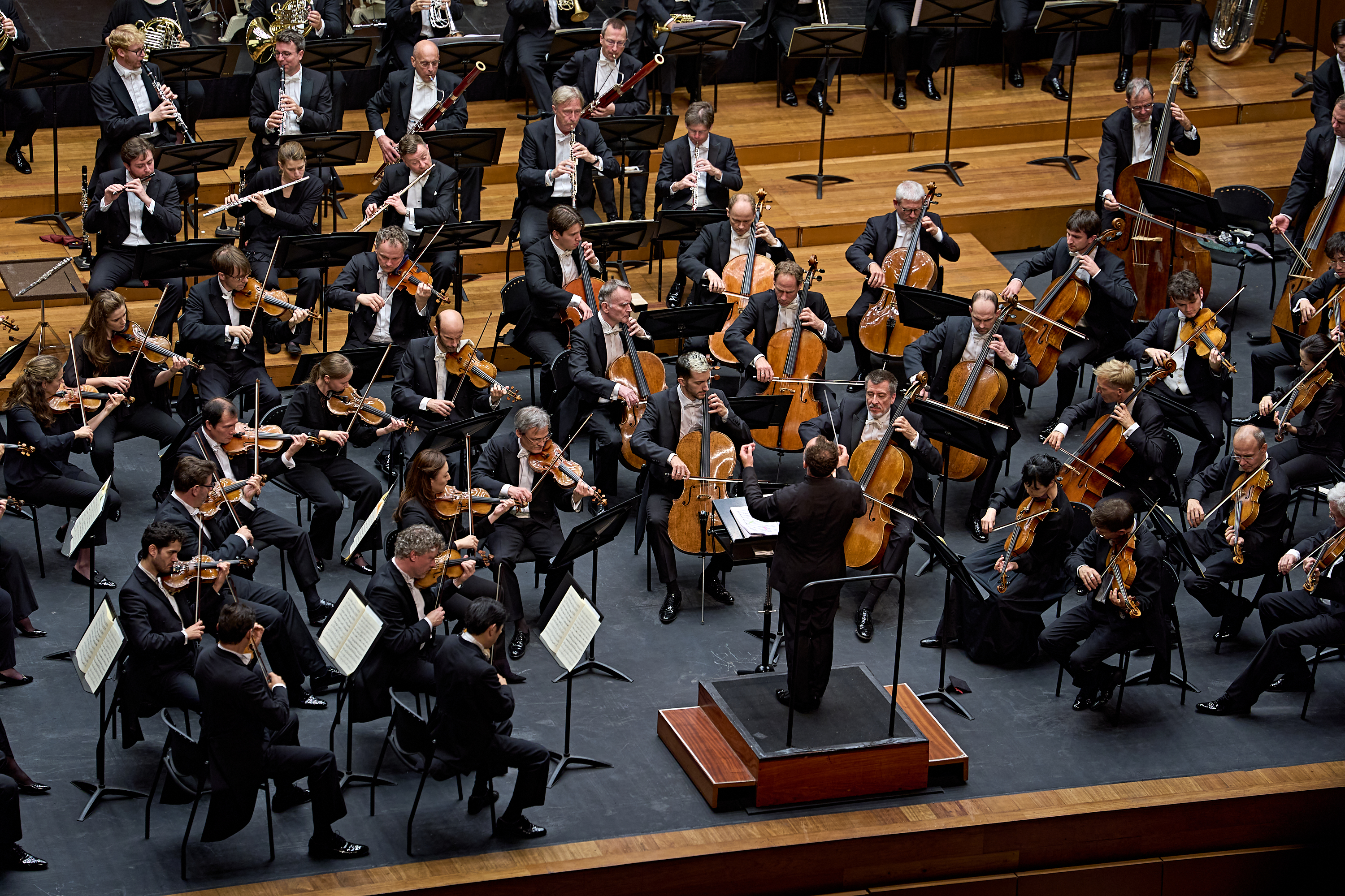 Ein vollbesetztes Sinfonieorchester, das auf der Bühne spielt, unter der Leitung eines Dirigenten, der auf einem Podium steht.