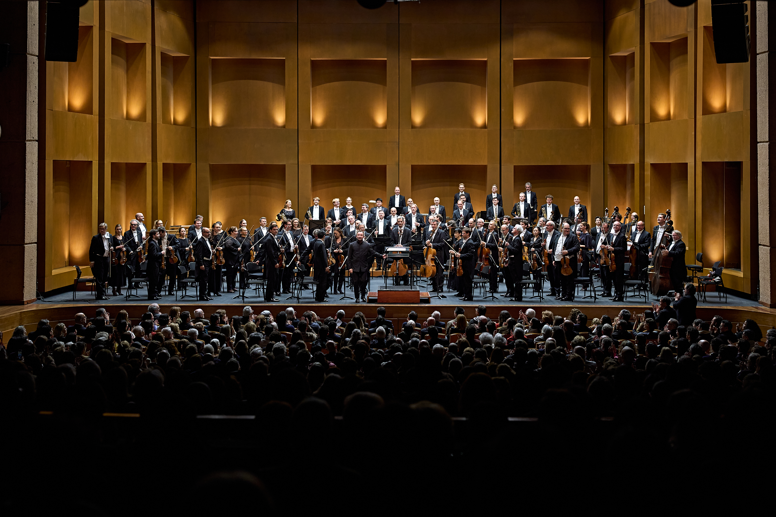 Ein vollbesetztes Orchester spielt auf einer Holzbühne vor einem sitzenden Publikum in einem Konzertsaal.