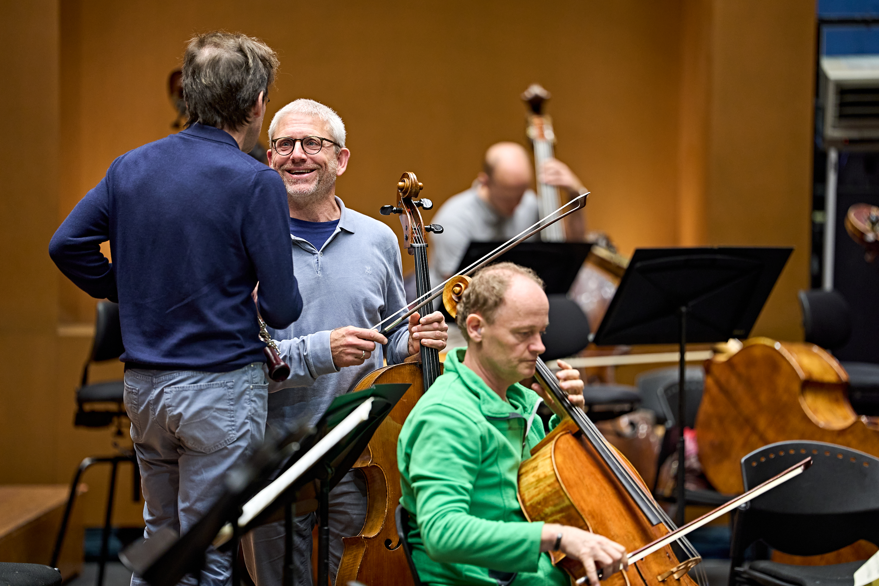 Zwei Musiker in einem Orchester, die ein Cello in der Hand halten, unterhalten sich, während im Vordergrund ein weiterer Cellist spielt und im Hintergrund ein Kontrabassist sitzt.