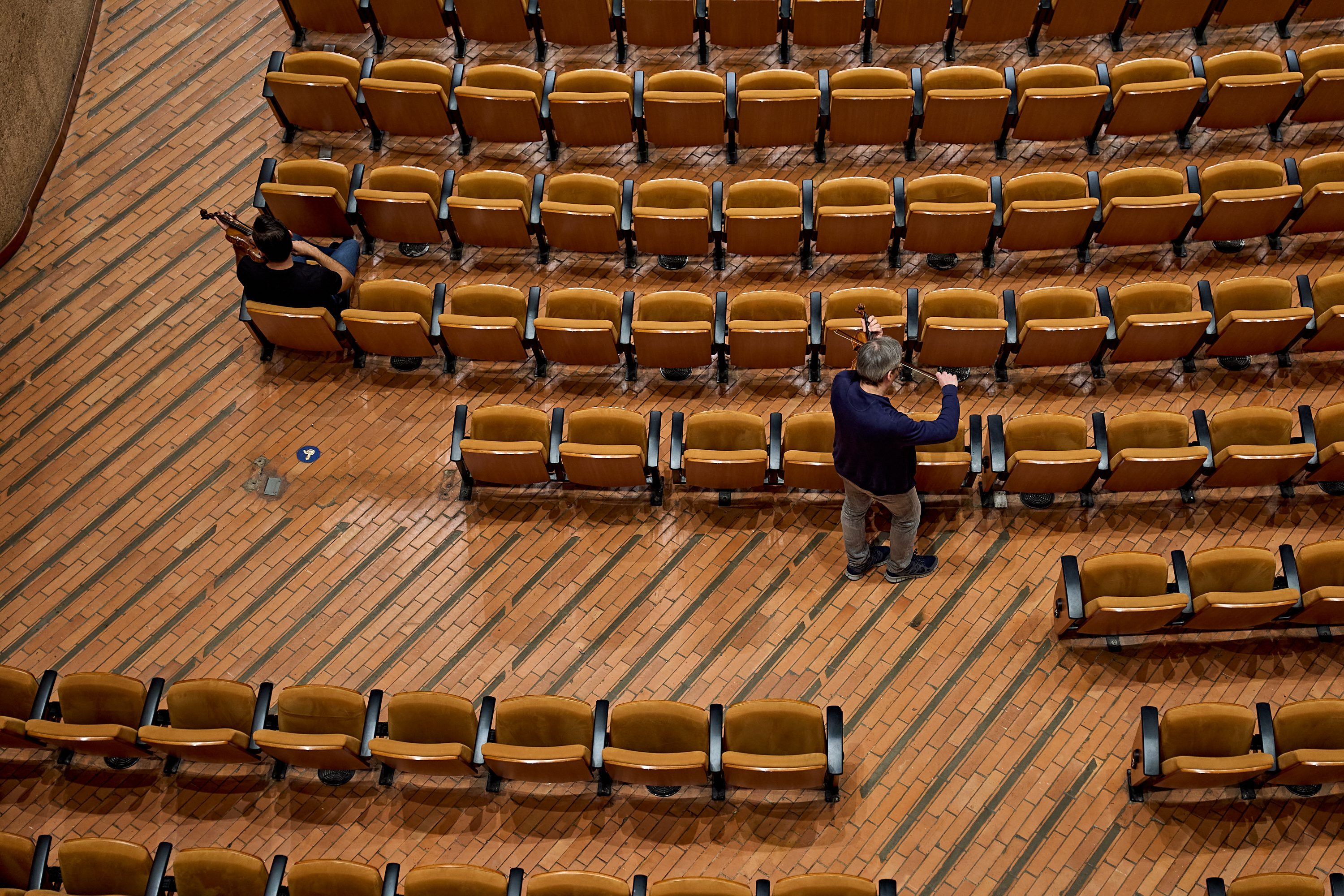Zwei Geiger üben in einem leeren Saal, der mit Reihen hellbrauner Stühle auf einem Ziegelboden ausgestattet ist.