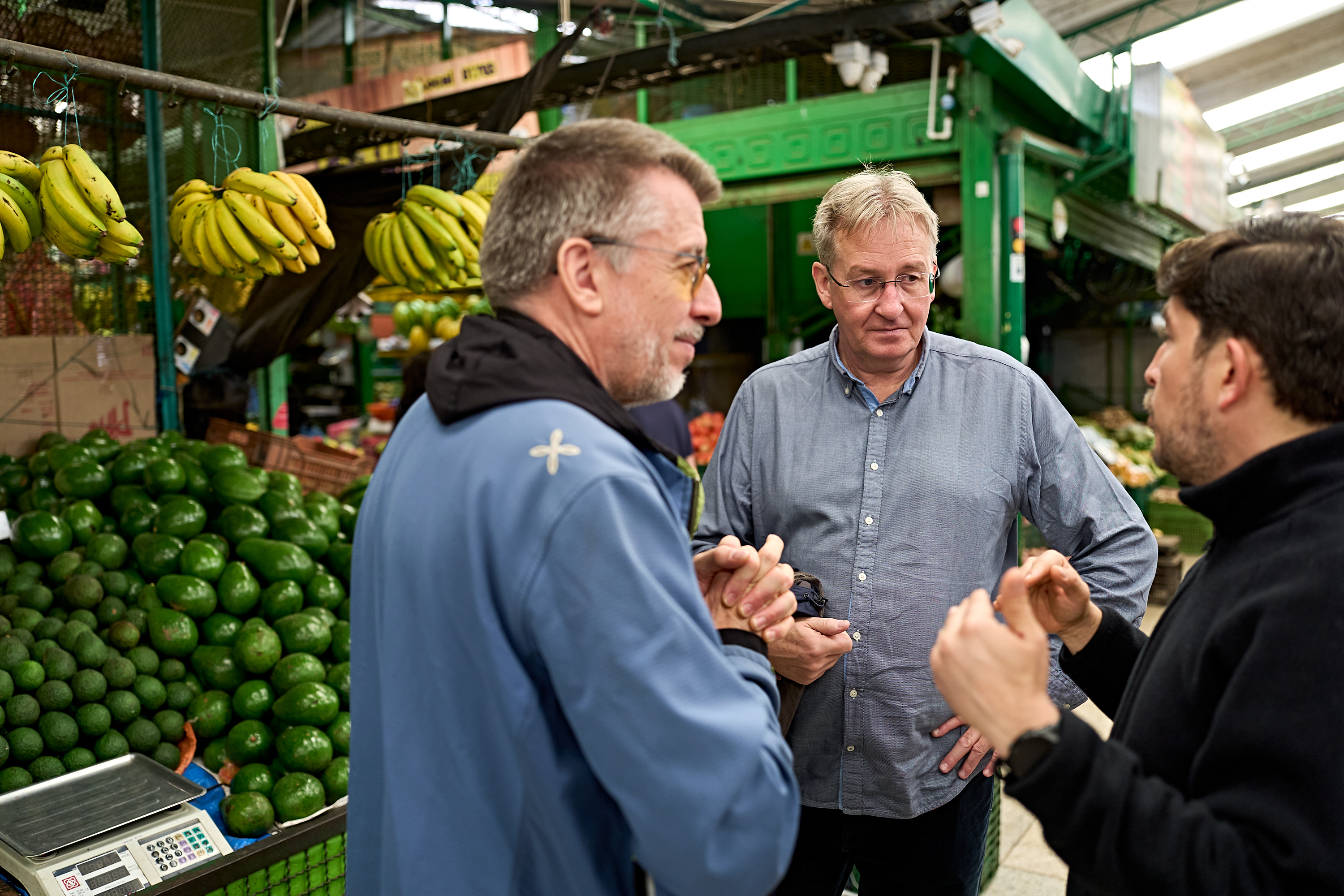 Drei Männer stehen zusammen und unterhalten sich an einem Marktstand, der mit Bananen- und Avocadobündeln gefüllt ist.