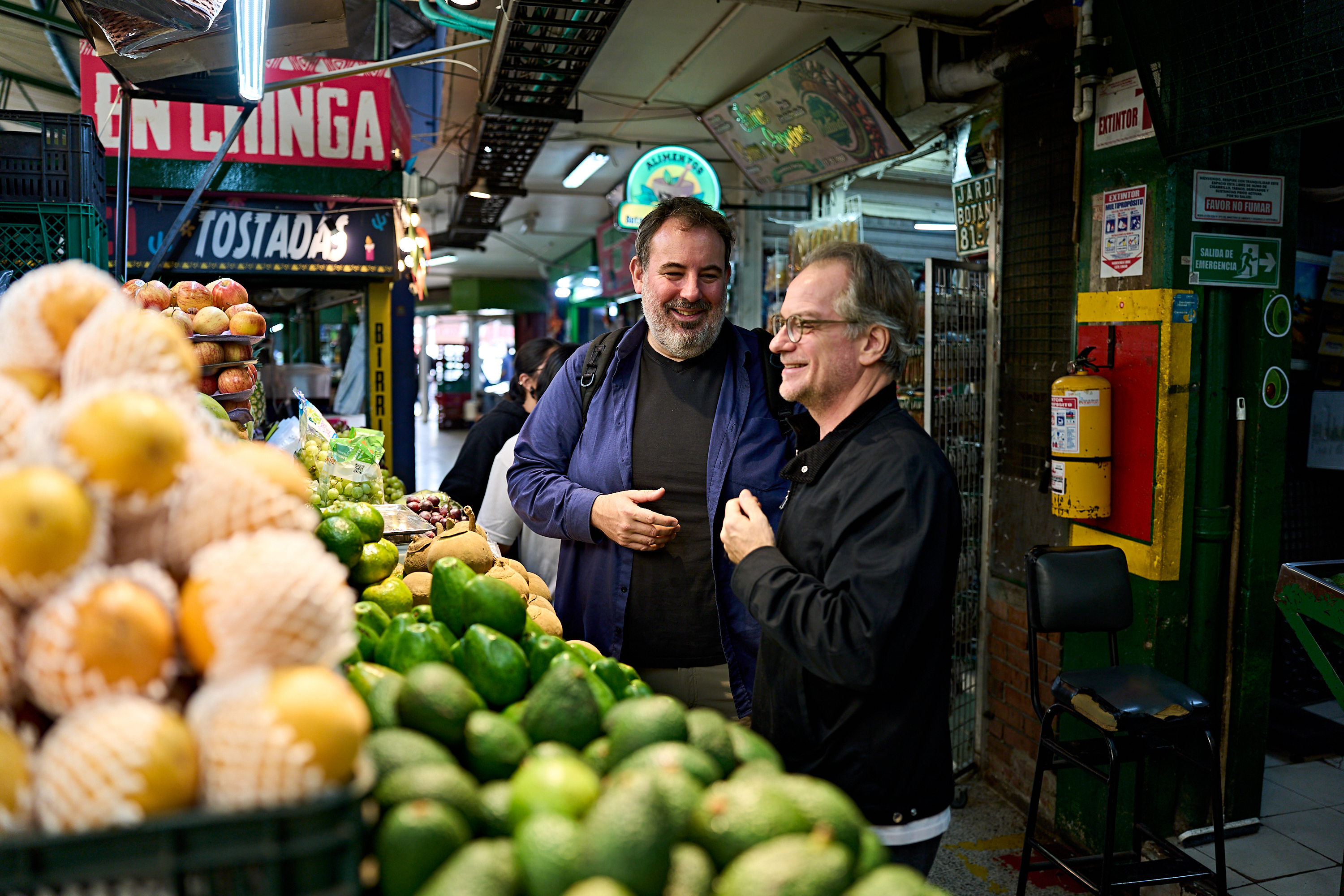 Zwei Männer stehen in einem mit Obst und Gemüse gefüllten Gang eines Supermarkts, lächeln und unterhalten sich miteinander.