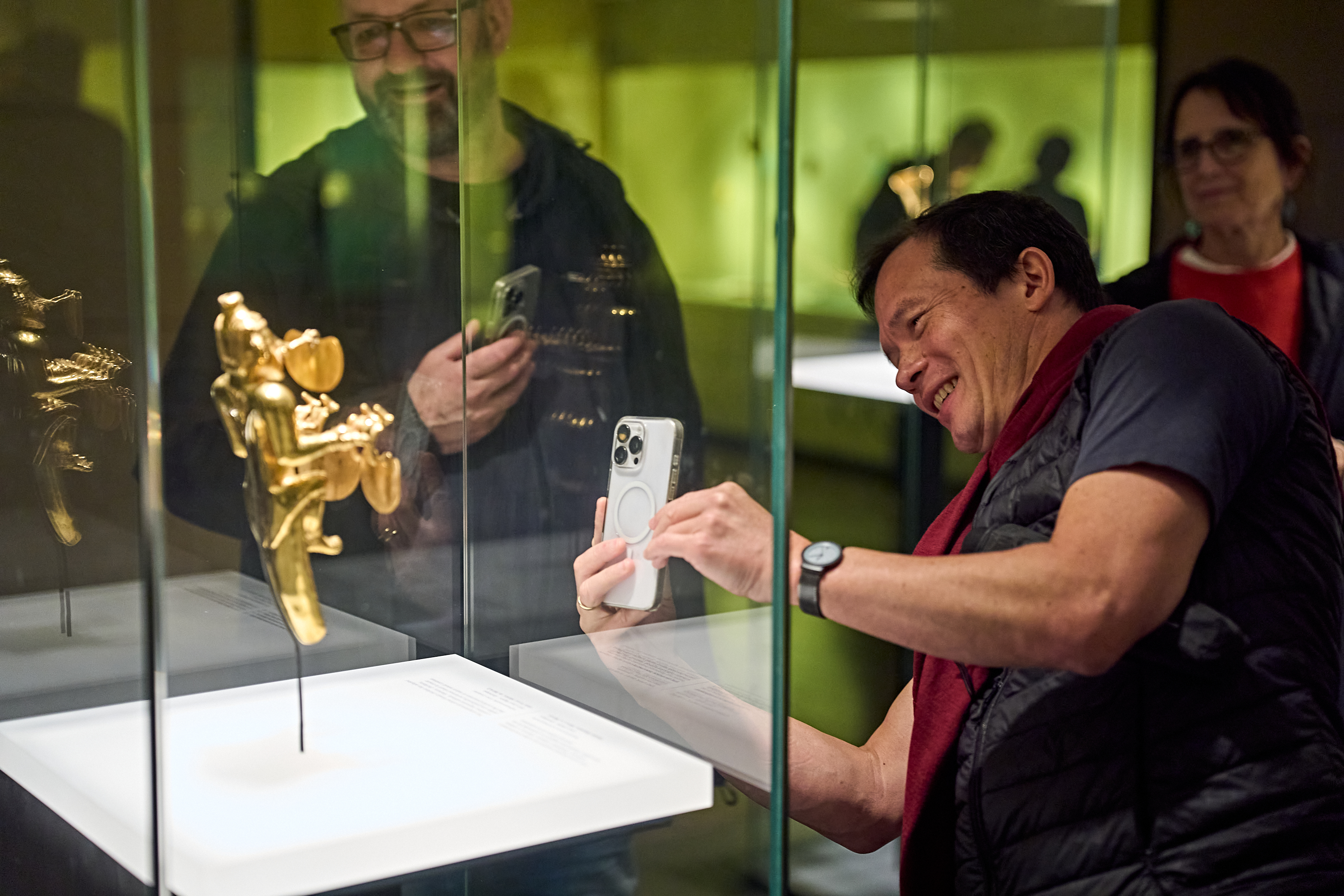 Ein Mann lächelt, während er mit seinem Handy eine kleine goldene Figur in einer Glasvitrine in einem Museum fotografiert.