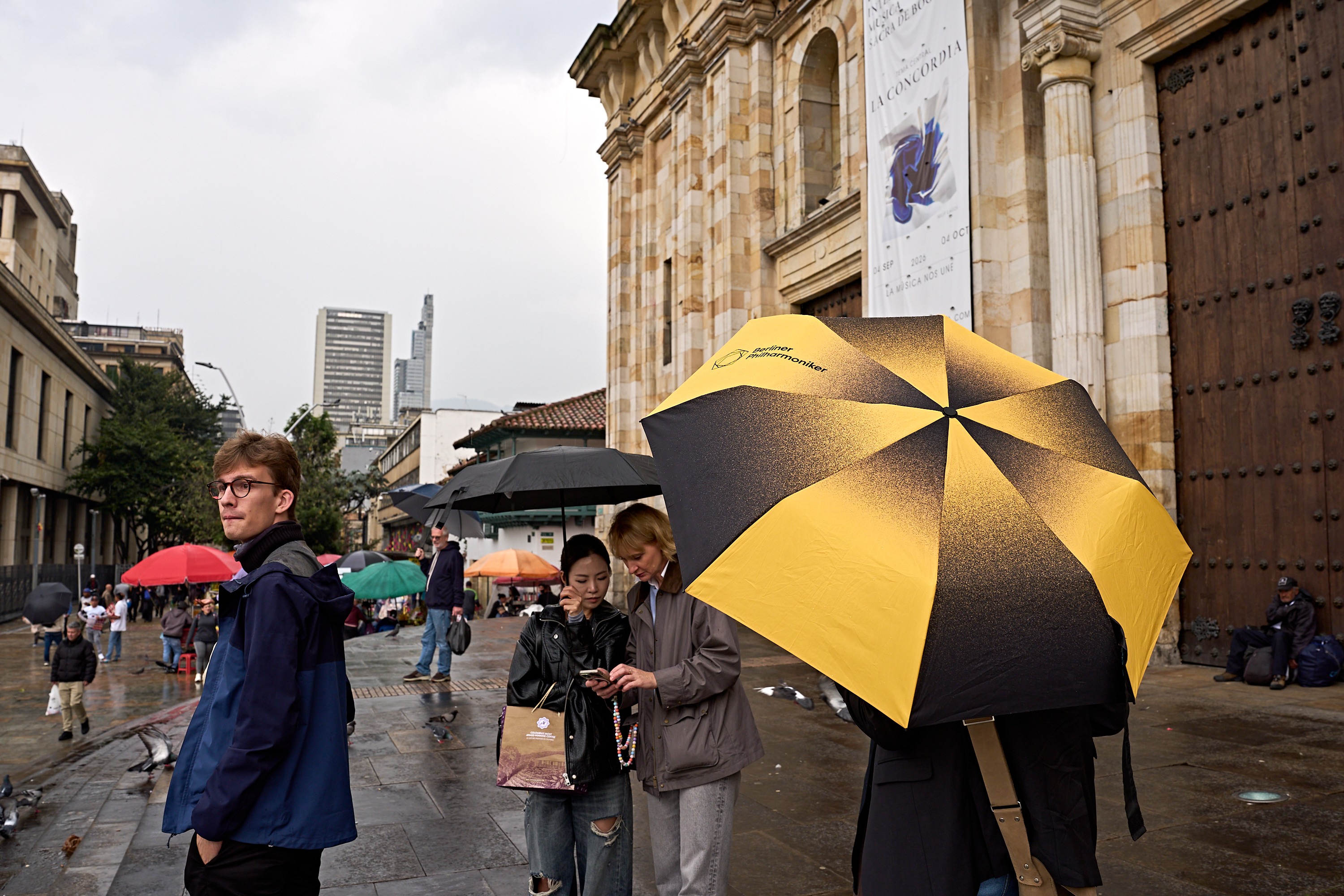 Menschen, die an einem regnerischen Tag über einen Platz gehen, im Vordergrund ein großer gelb-schwarzer Regenschirm der Berliner Philharmoniker.