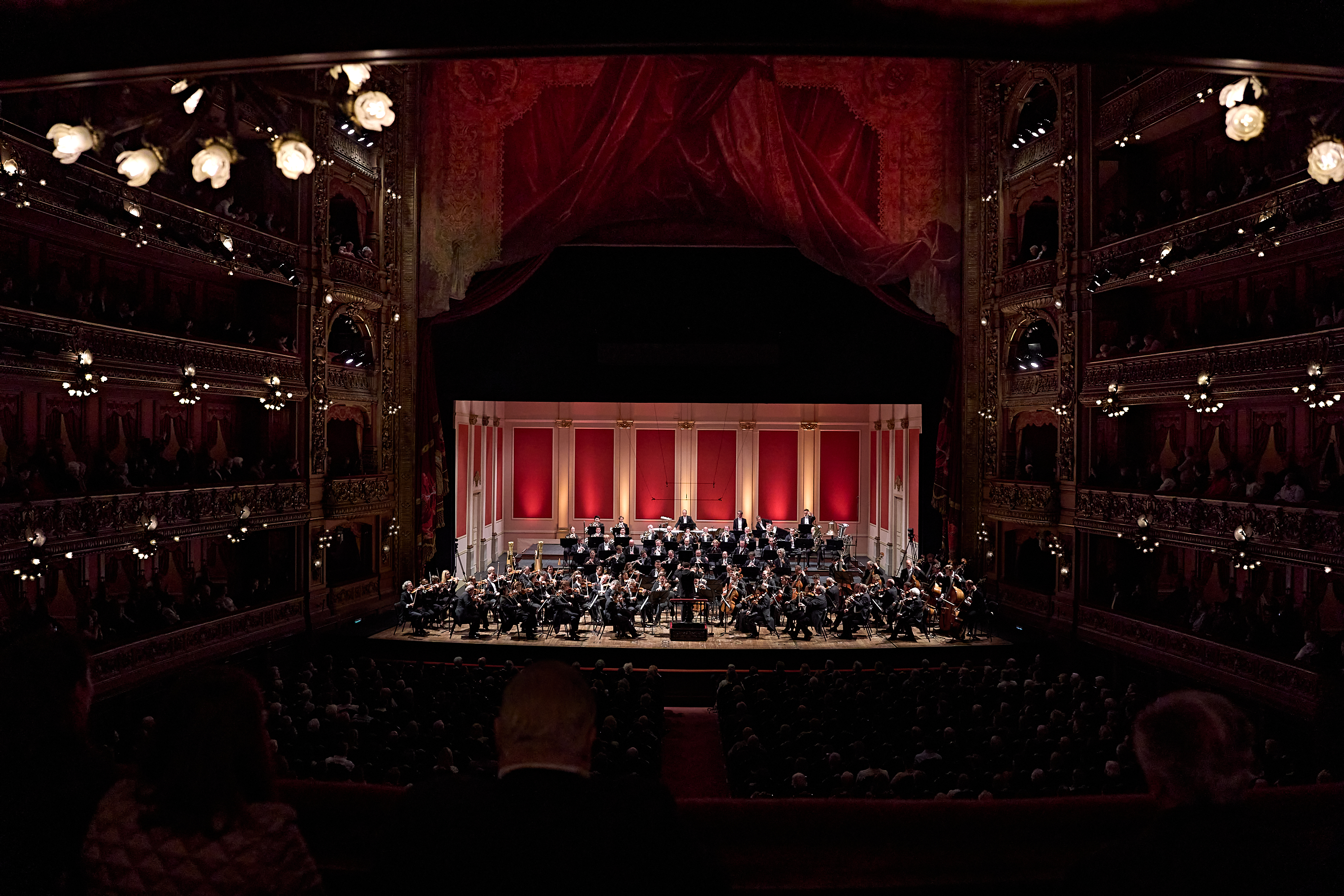 Ein Orchester spielt auf der Bühne eines prächtigen, mehrstöckigen Theaters, das vom Balkon aus zu sehen ist.