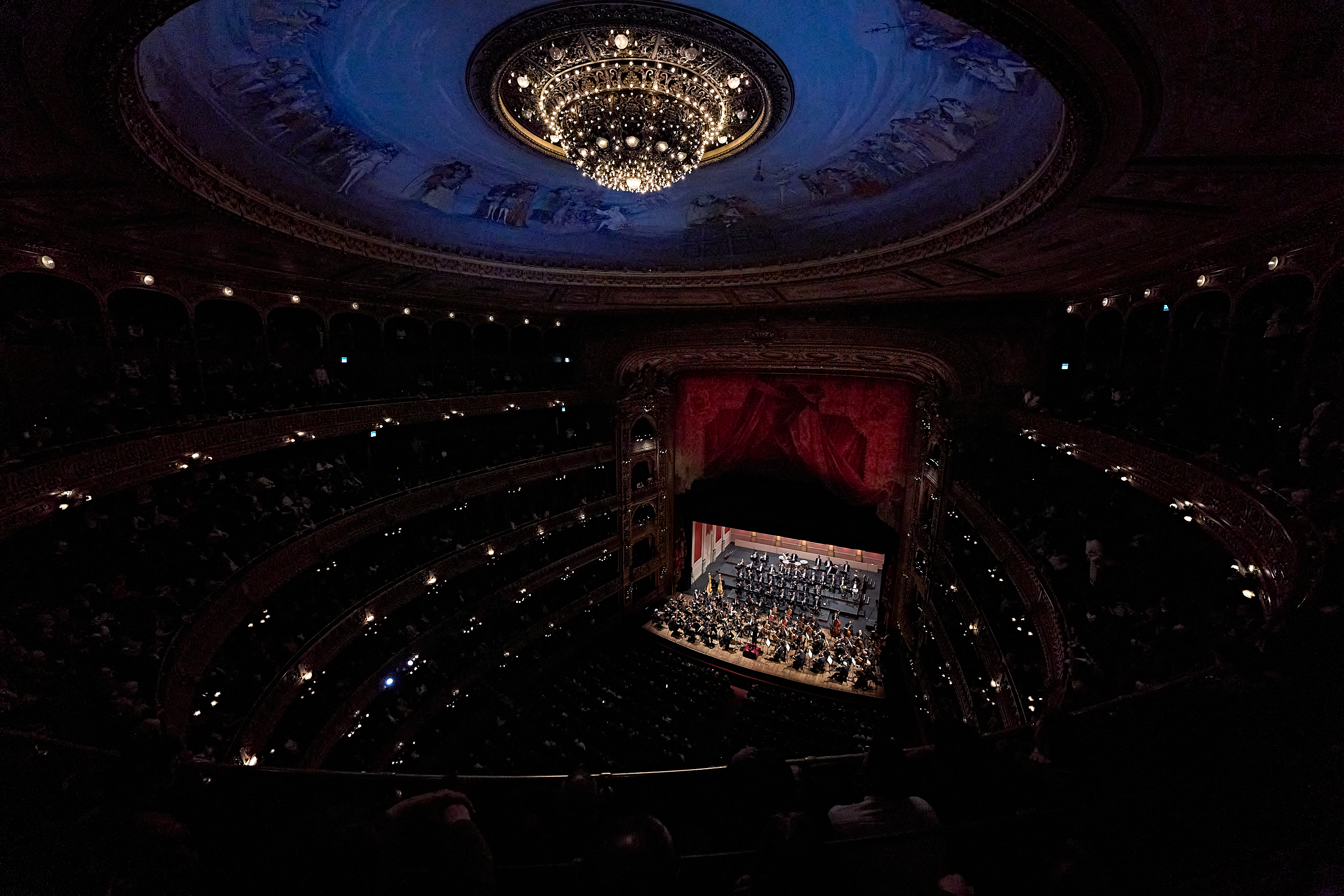 Eine Aufnahme aus großer Höhe eines prächtigen, mehrstöckigen Theaters, in dem ein Orchester auf der Bühne unter einem großen, kunstvoll verzierten Kronleuchter und einem gemalten Deckenfresko spielt.