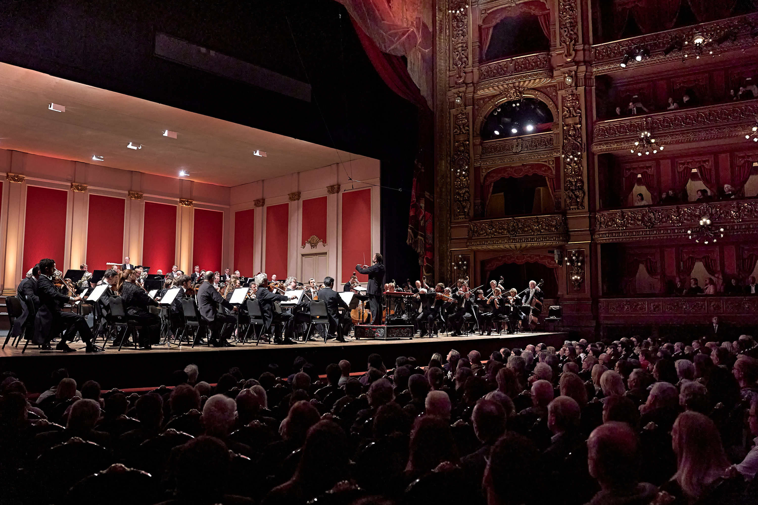 Ein vollbesetztes Orchester spielt auf der Bühne eines prunkvollen, mehrstöckigen Theaters, während das Publikum von den Rängen aus zusieht.
