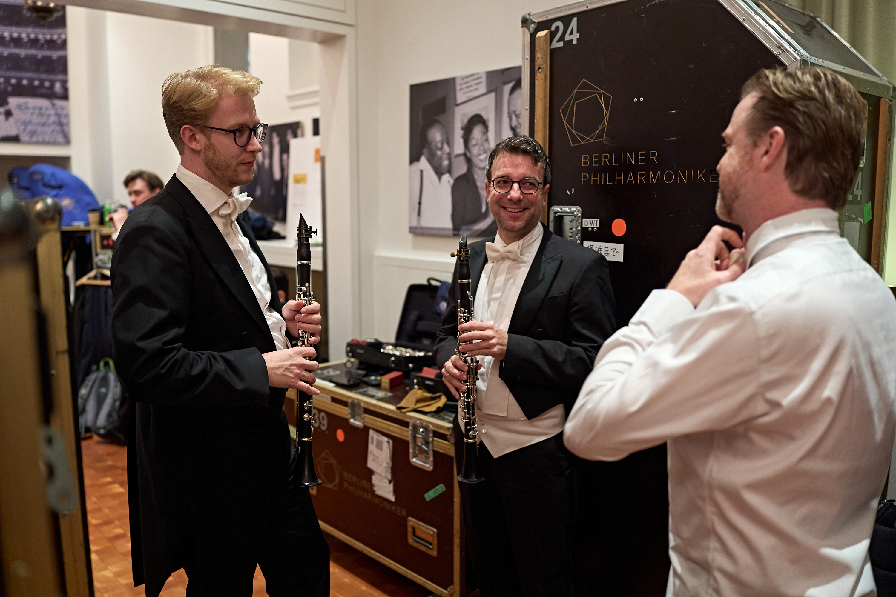 Zwei Männer in eleganten Smokings halten Klarinetten in den Händen, während ein dritter Mann in einem weißen Hemd hinter der Bühne der Berliner Philharmoniker seine Fliege zurechtzieht.