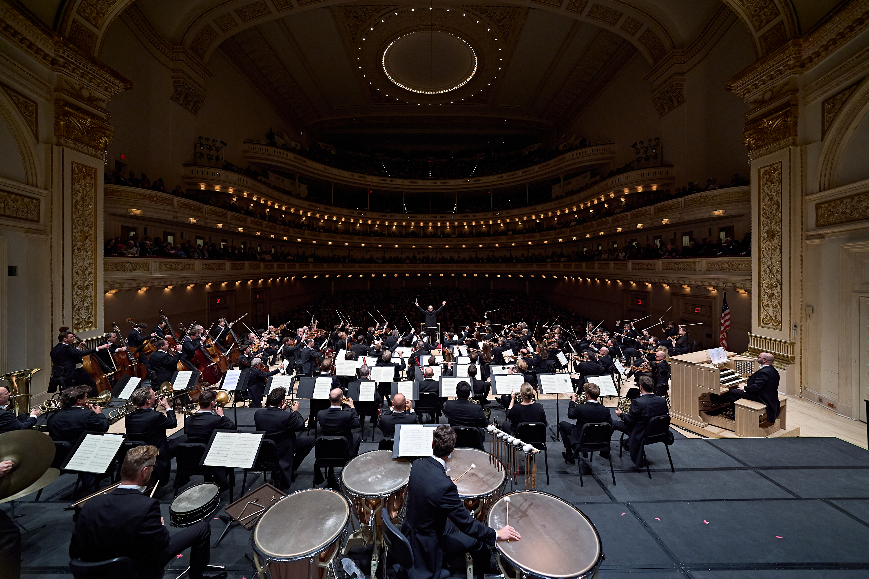 Ein Orchester spielt auf der Bühne eines prächtigen, mehrstöckigen Konzertsaals; die Aufnahme zeigt den Blick von hinter der Schlagzeuggruppe.