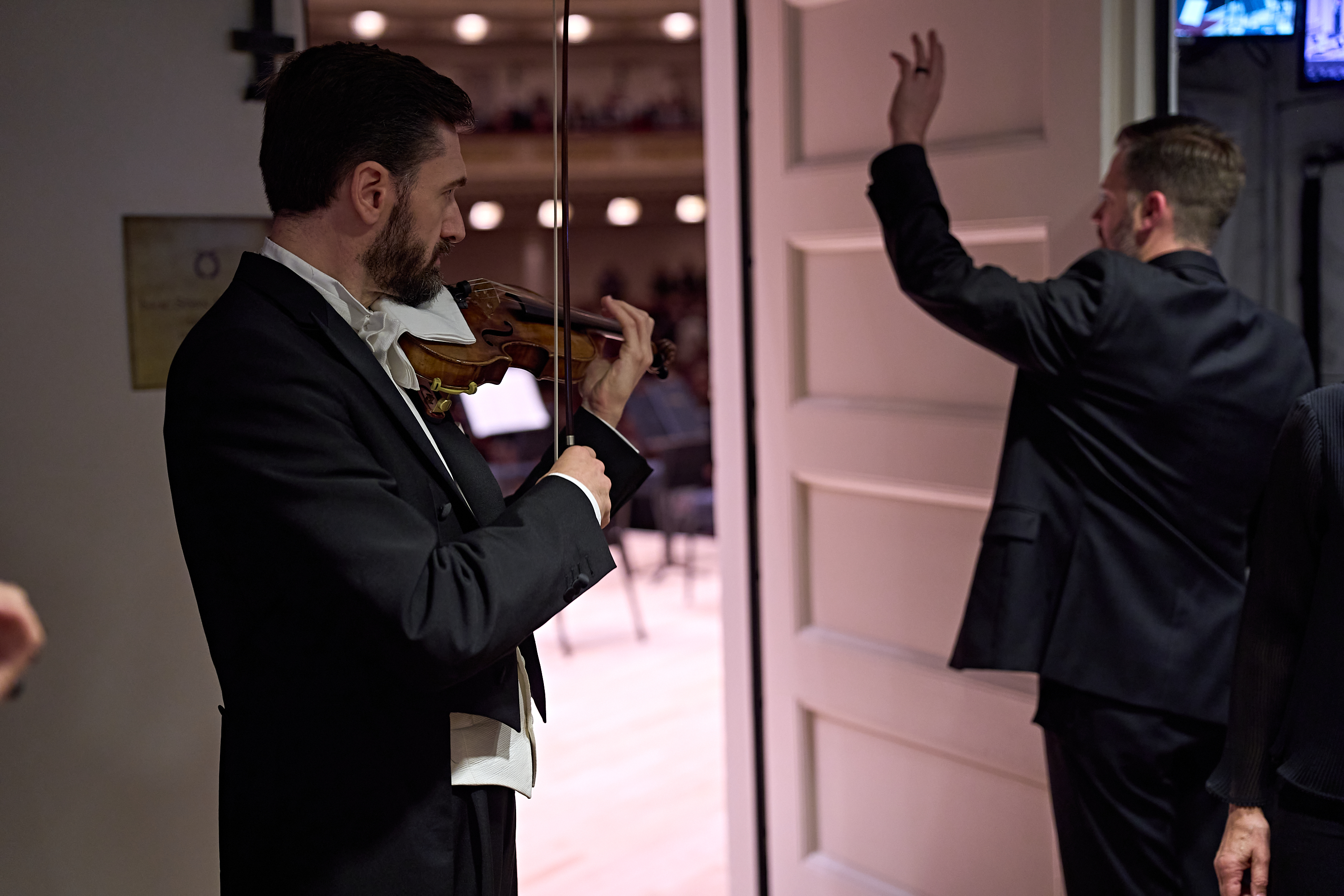 A violinist in a tuxedo plays his instrument while a staff member gives a hand signal at the stage entrance.