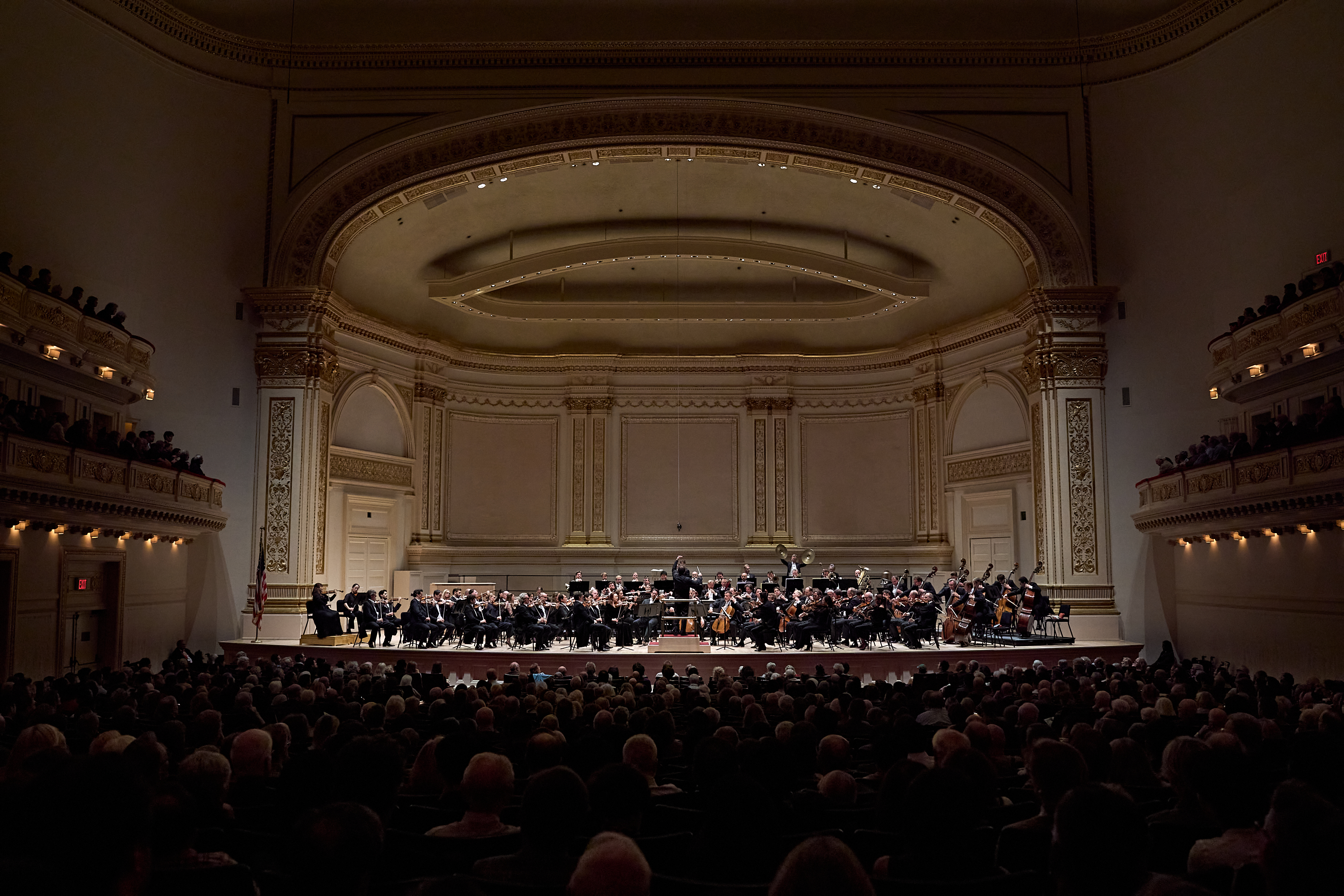 Ein Orchester spielt auf der Bühne in einem prächtigen, mehrstöckigen Konzertsaal, der mit Zuschauern gefüllt ist.