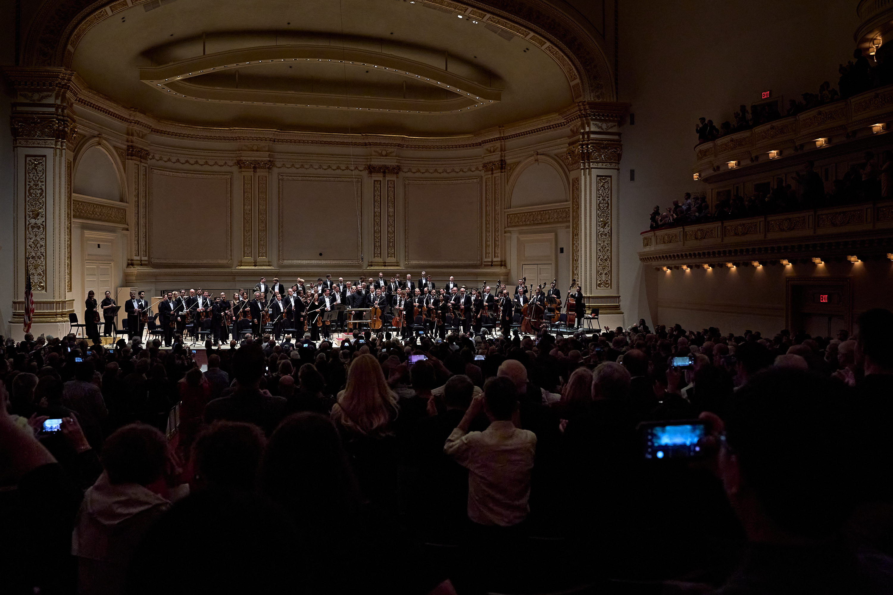 Die Musiker*innen eines Orchesters stehen nach dem Konzert auf der Bühne eines prächtigen Konzertsaals und nehmen den Applaus entgegen, während Zuschauer*innen im vollbesetzten Saal mit ihren Handys Fotos machen.