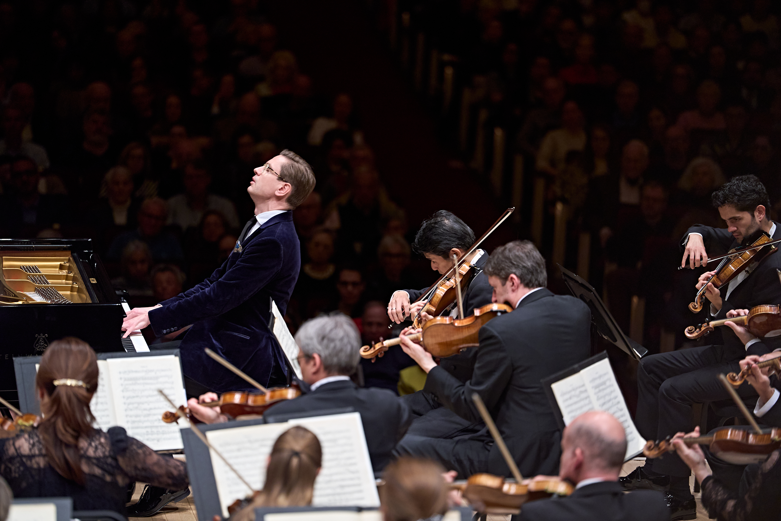 Ein Pianist in einer dunklen Samtjacke spielt auf einem Flügel und dirigiert dabei ein Orchester auf der Bühne vor Publikum.