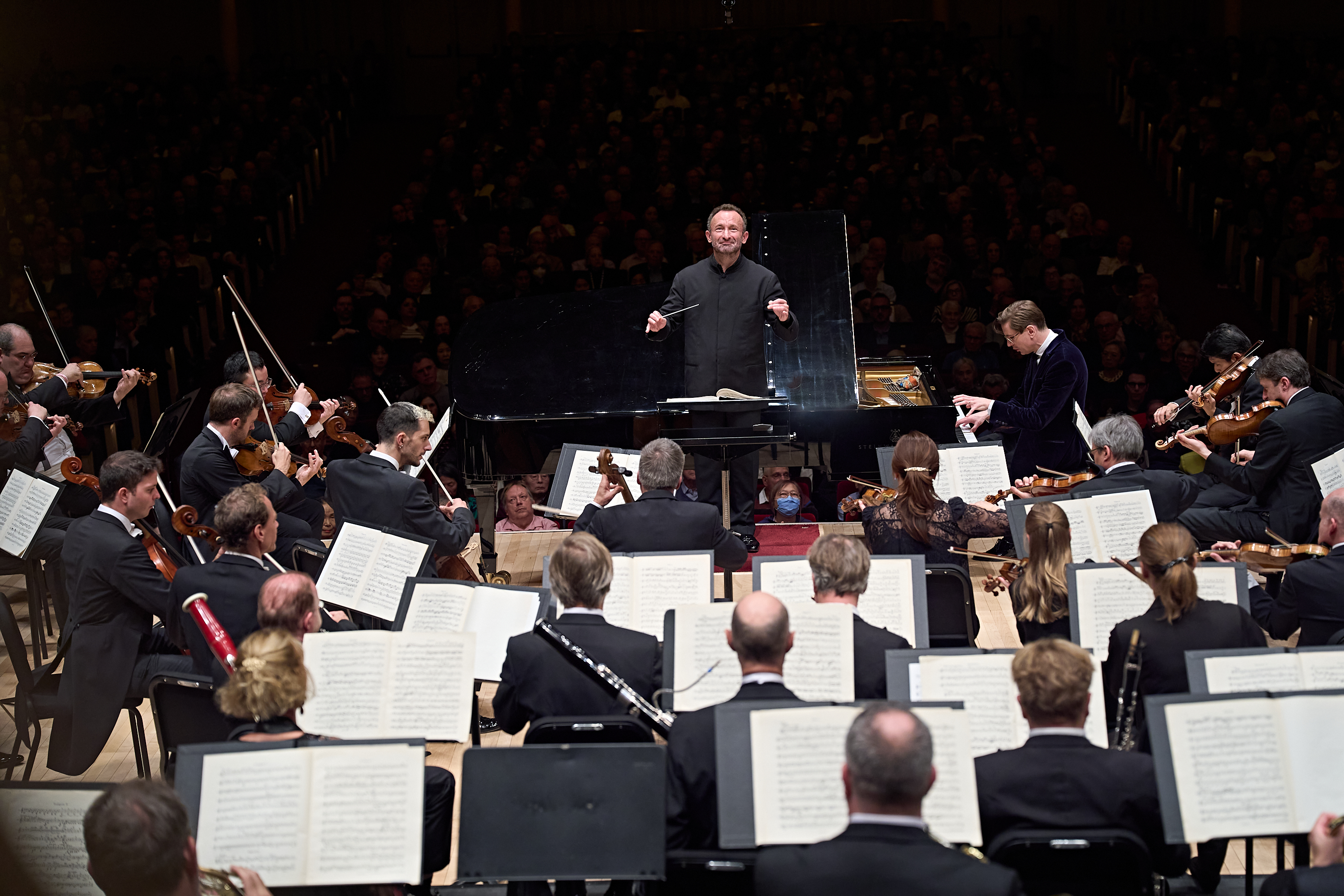 Ein Dirigent leitet ein Orchester auf der Bühne, während ein Pianist am Flügel spielt und im Hintergrund ein großes Publikum sitzt.