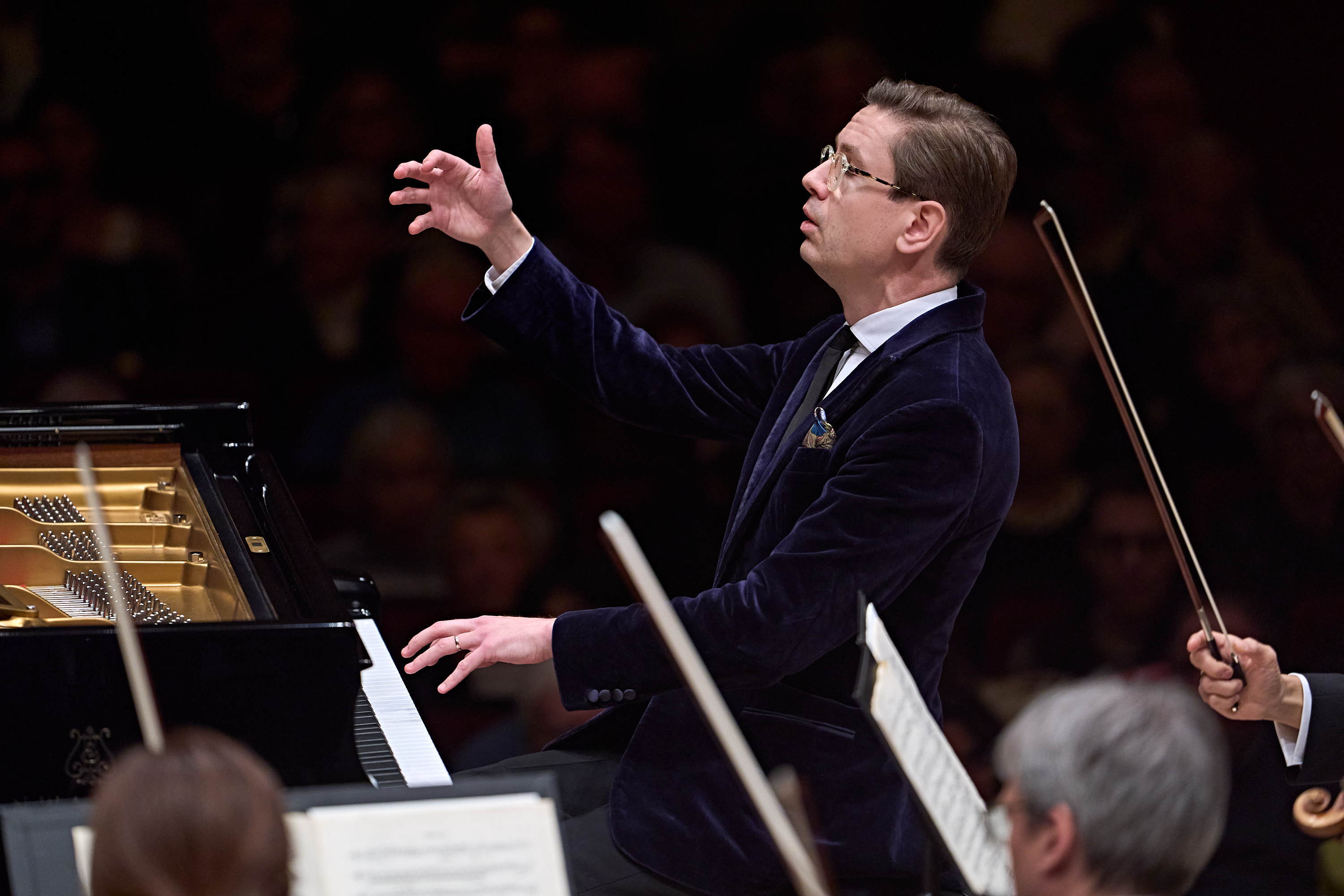 Ein Pianist in einem dunklen Samtanzug sitzt am Klavier und spielt, ein Orchester begleitet ihn.