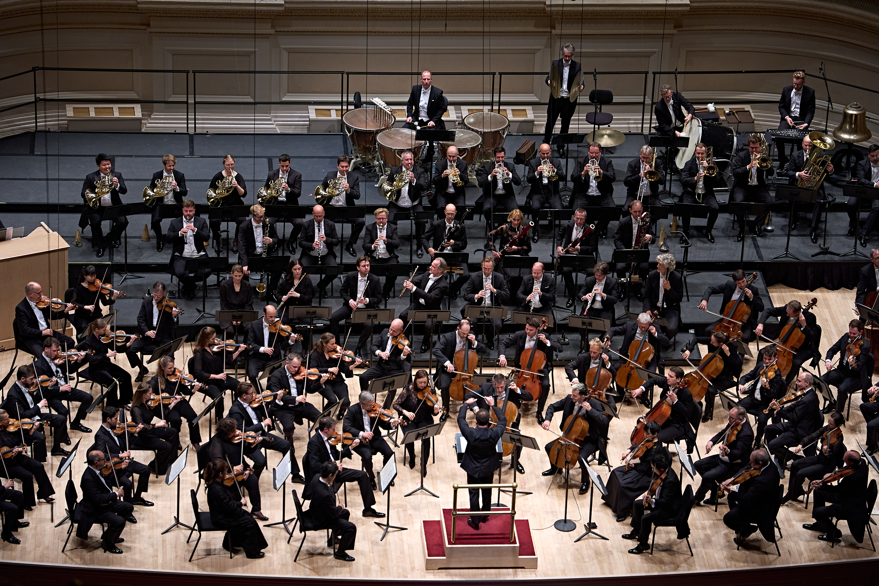 Ein vollbesetztes Orchester, das auf einer Bühne spielt, unter der Leitung eines Dirigenten, der auf einem Podium steht.