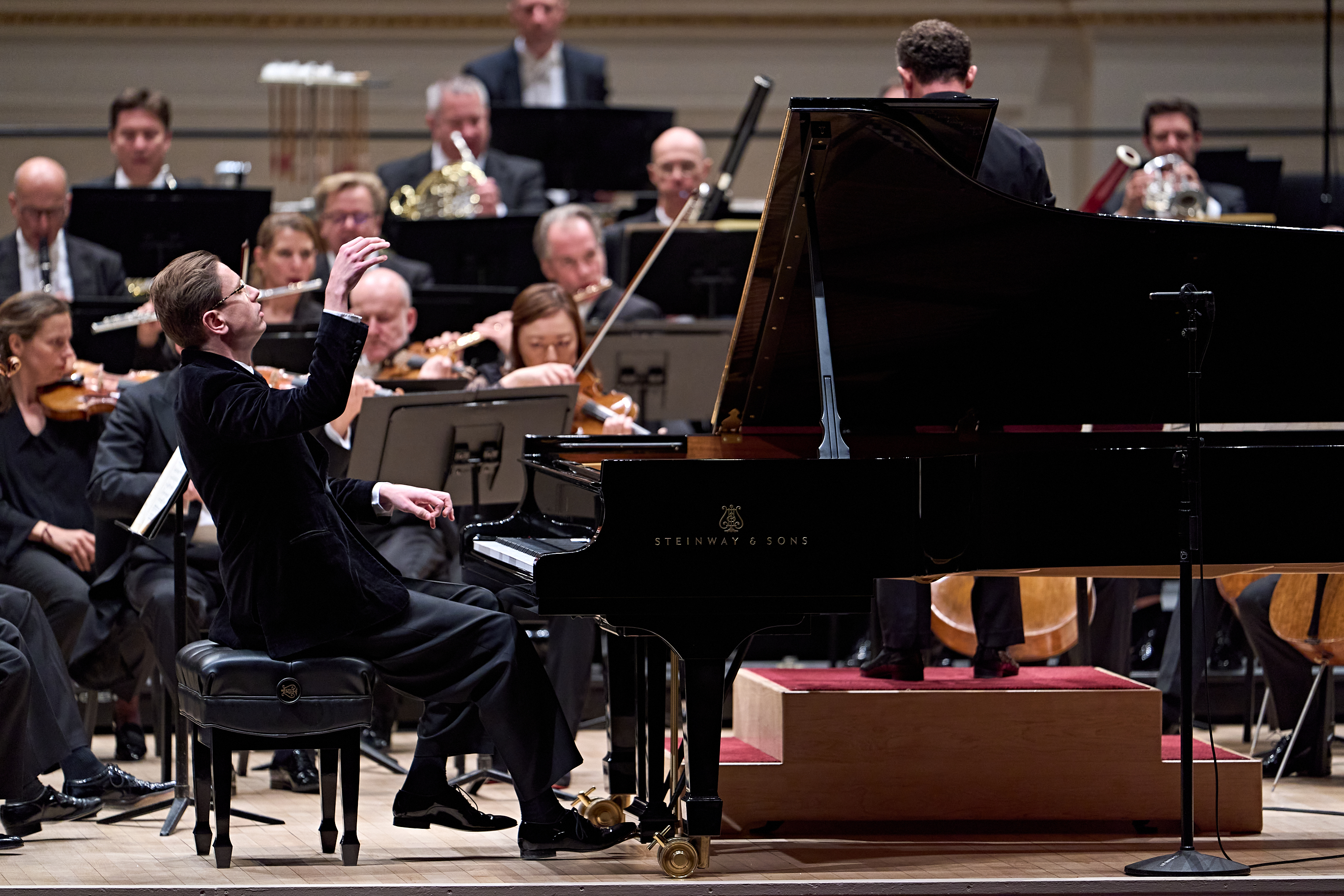 Ein Pianist in einem dunklen Anzug spielt auf einem Flügel, umgeben von einem Orchester auf einer Konzertbühne.