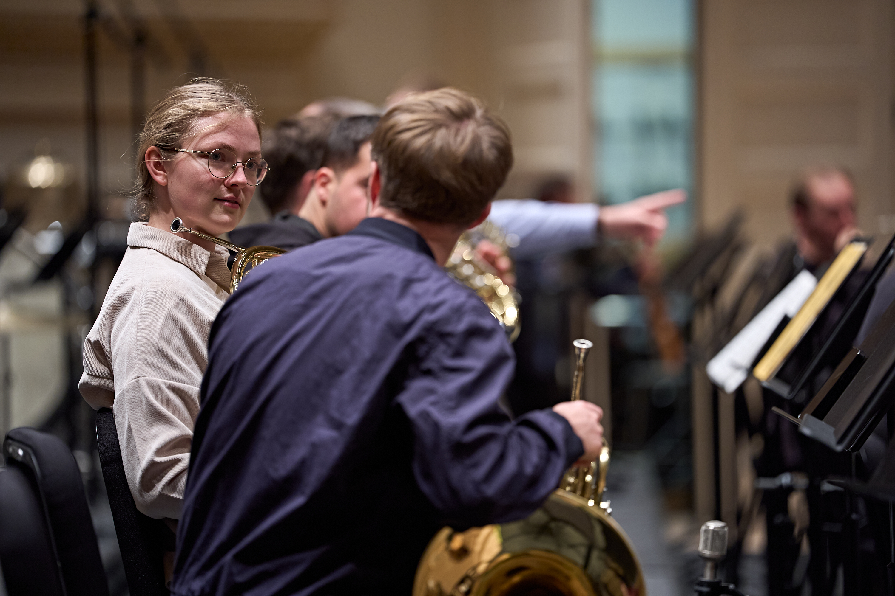 Zwei Hornisten sitzen in einem Orchester, einer blickt in die Kamera, während der andere nach vorne schaut.