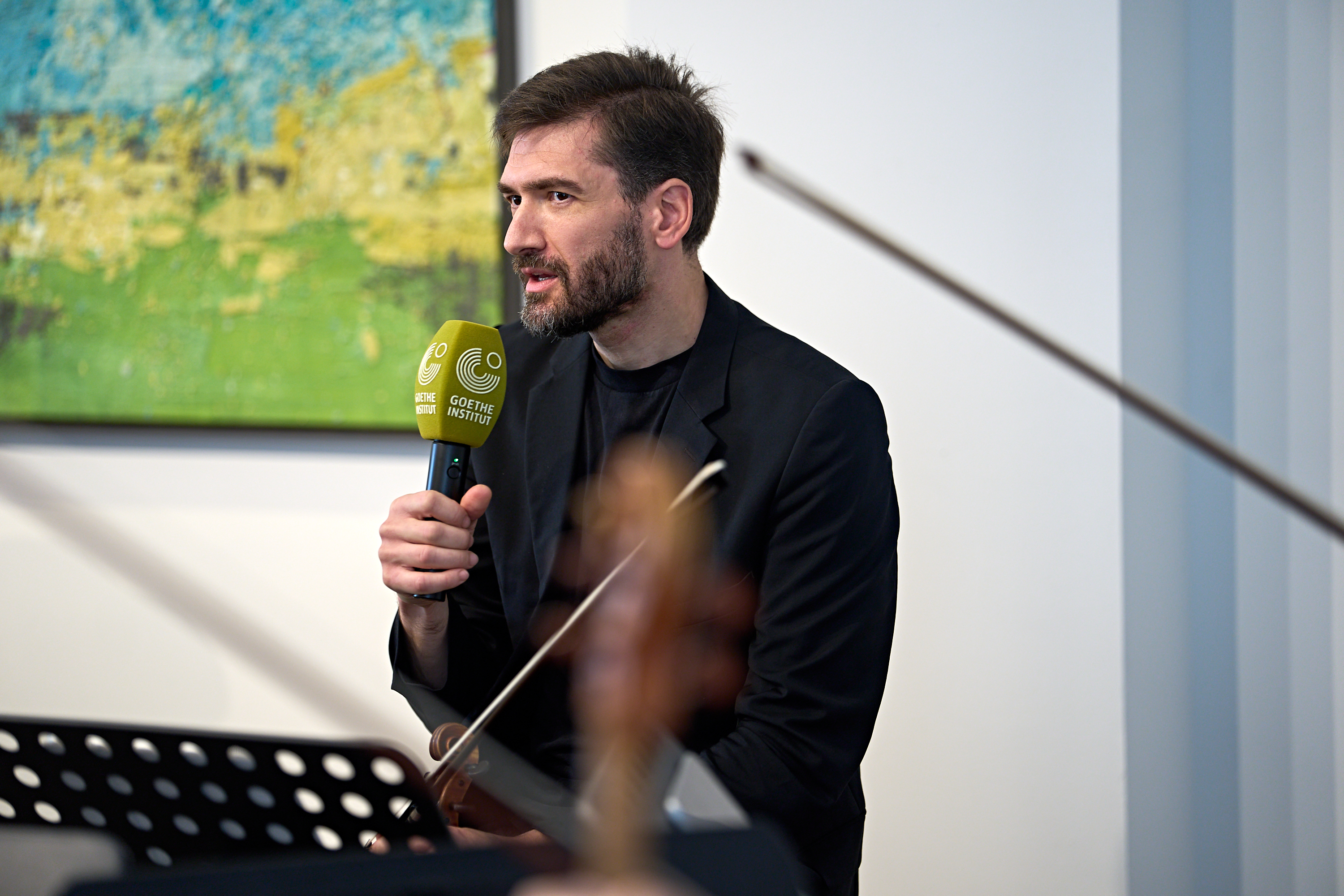 Ein Mann in einem schwarzen Blazer spricht während einer Musikaufführung in ein Mikrofon, auf dem das Logo des Goethe-Instituts zu sehen ist.