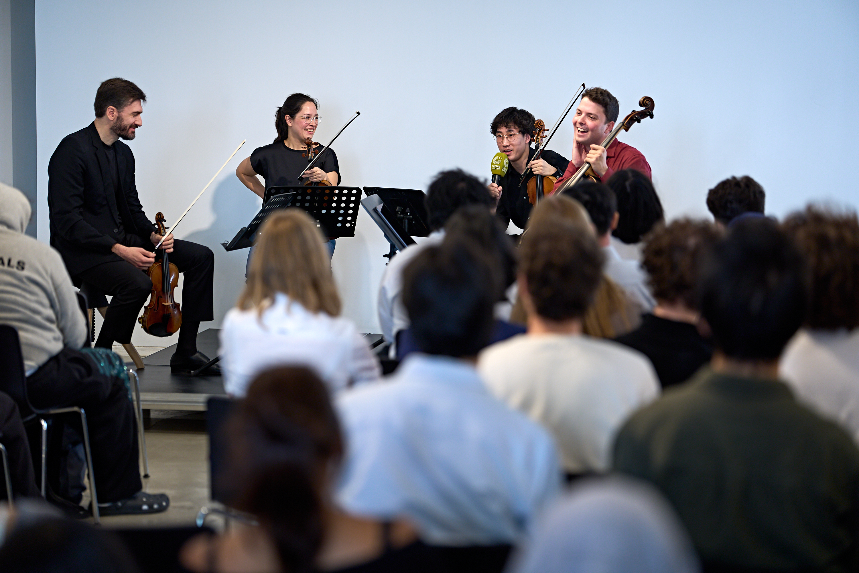 Drei Musiker treten auf der Bühne mit Saiteninstrumenten auf und sprechen dabei vor Publikum in ein Mikrofon.