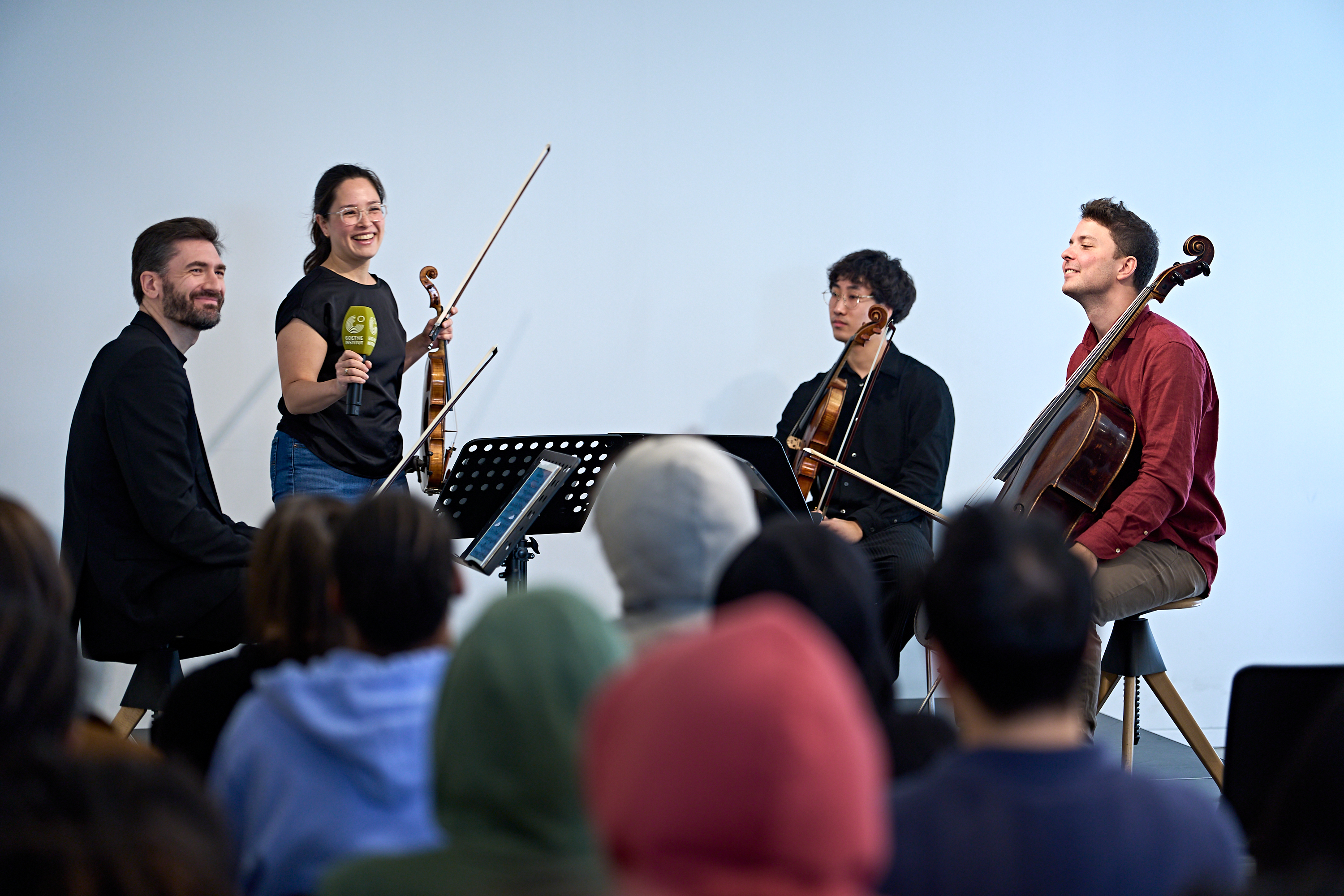 Eine Frau, die eine Geige und ein Mikrofon in der Hand hält, spricht zu den Zuhörern, während zwei weitere Musiker in ihrer Nähe sitzen und eine Geige bzw. ein Cello halten.