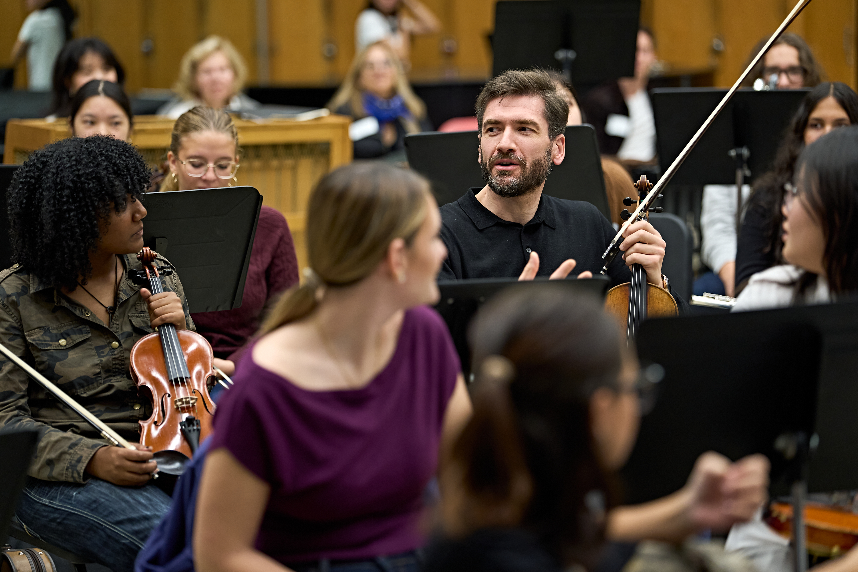 Ein Mann in einem schwarzen Hemd spricht während einer Orchesterprobe mit einer Gruppe von Geigern.