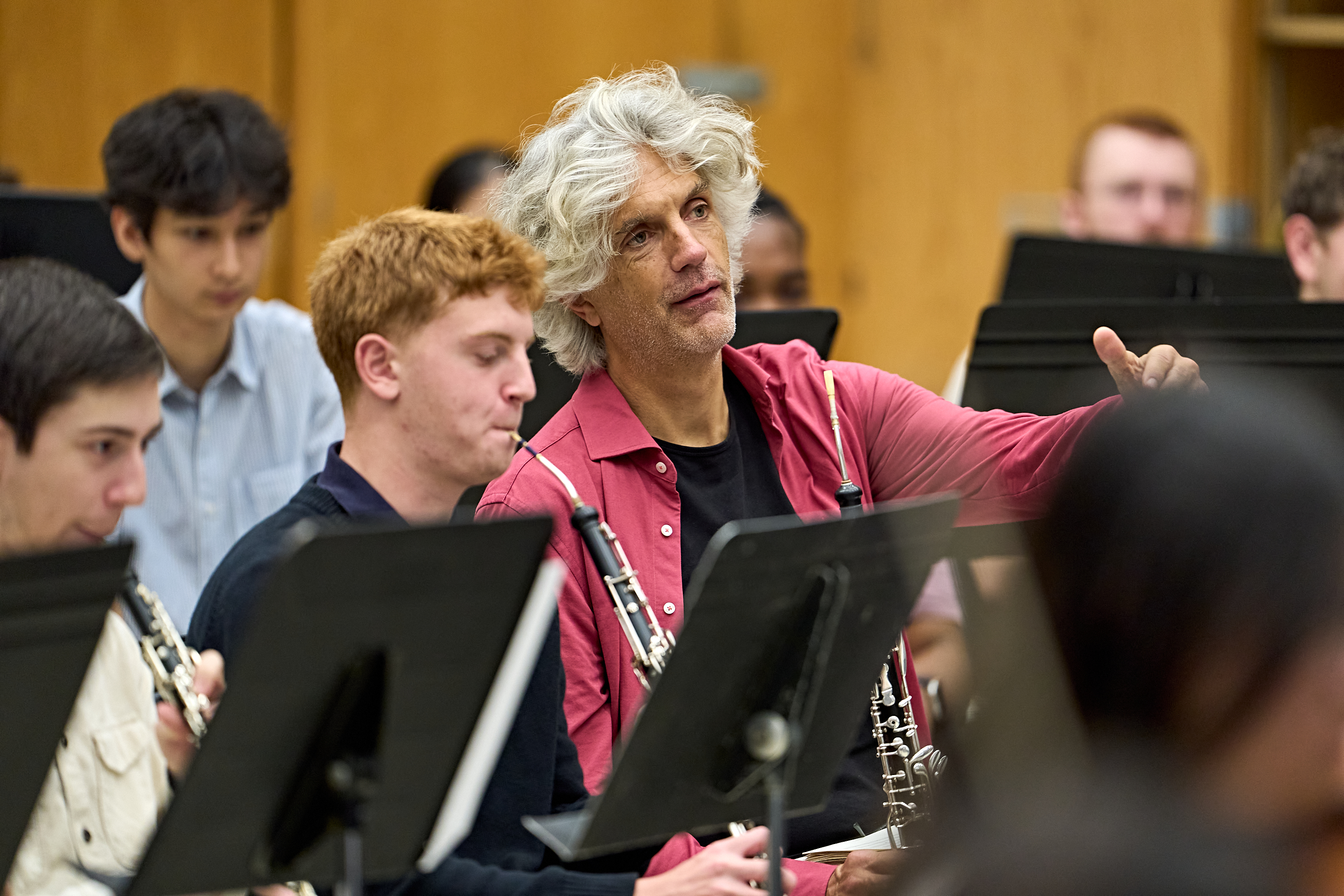 Ein Mann mit grauen Haaren in einem roten Hemd gibt mit Gesten Anweisungen, während er ein Orchester aus Schülern unterrichtet, die Holzblasinstrumente spielen.