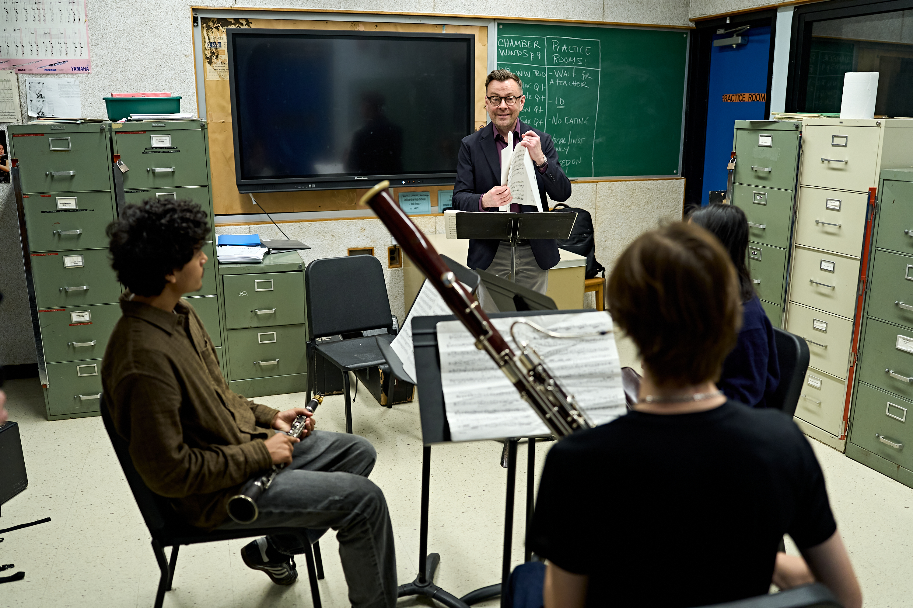 Ein Musiklehrer hält Notenblätter in der Hand, während er drei Schülern, die Klarinette und Fagott spielen, im Klassenzimmer Anweisungen gibt.