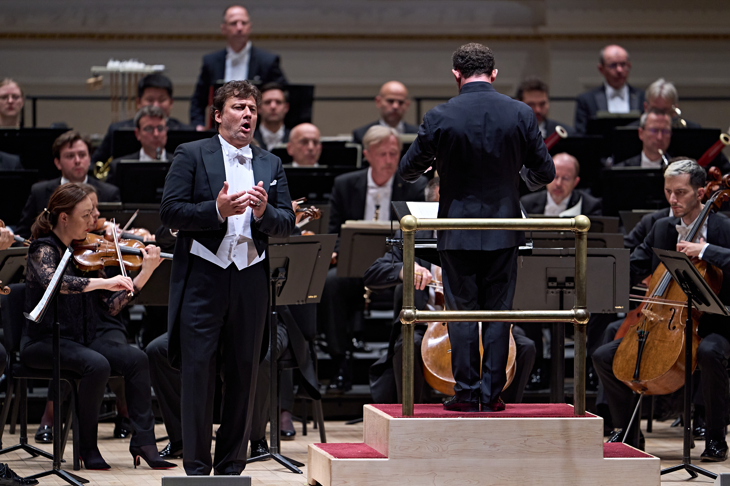 Ein männlicher Opernsänger im Smoking steht auf der Bühne und singt, hinter ihm steht ein Dirigent, der dem Orchester zugewandt ist.