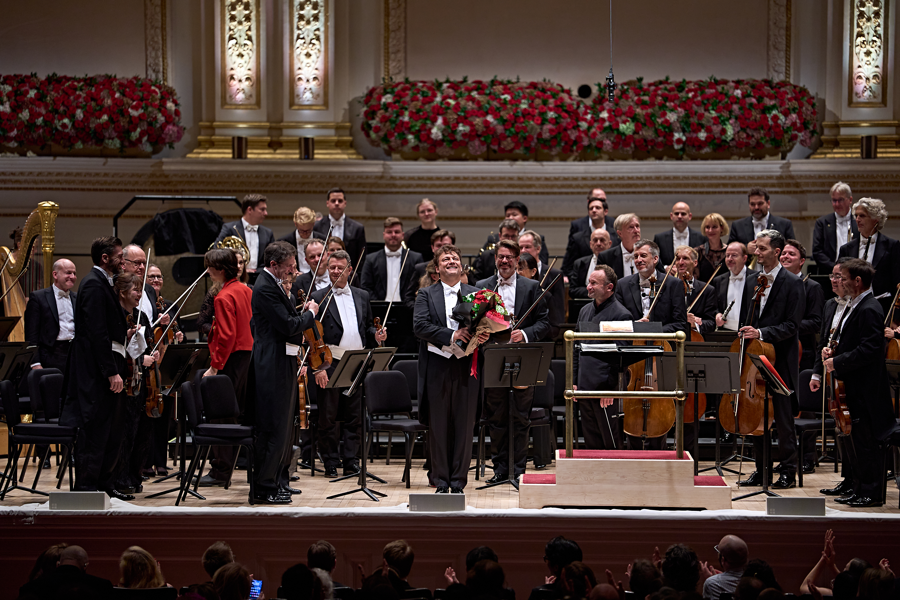 Ein Geiger, der einen Blumenstrauß in der Hand hält, steht in der Mitte der Bühne vor einem Orchester in der Carnegie Hall, während die Musiker und das Publikum applaudieren.