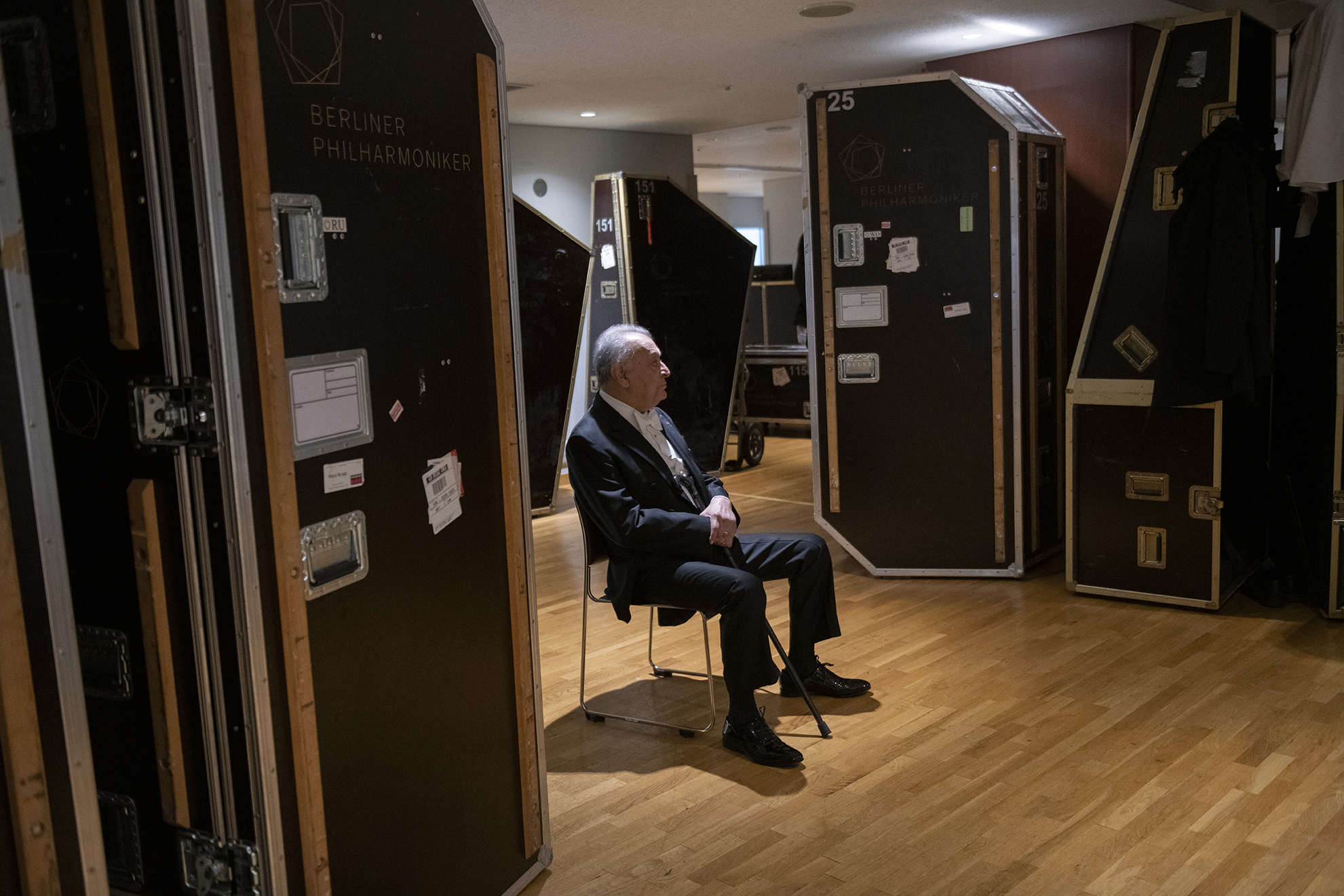 Ein älterer Mann im Anzug sitzt allein auf einem Stuhl in einem Backstage-Bereich, umgeben von hohen Instrumentenkästen mit der Aufschrift Berliner Philharmoniker. Das Licht ist gedämpft und der Boden ist aus Holz.
