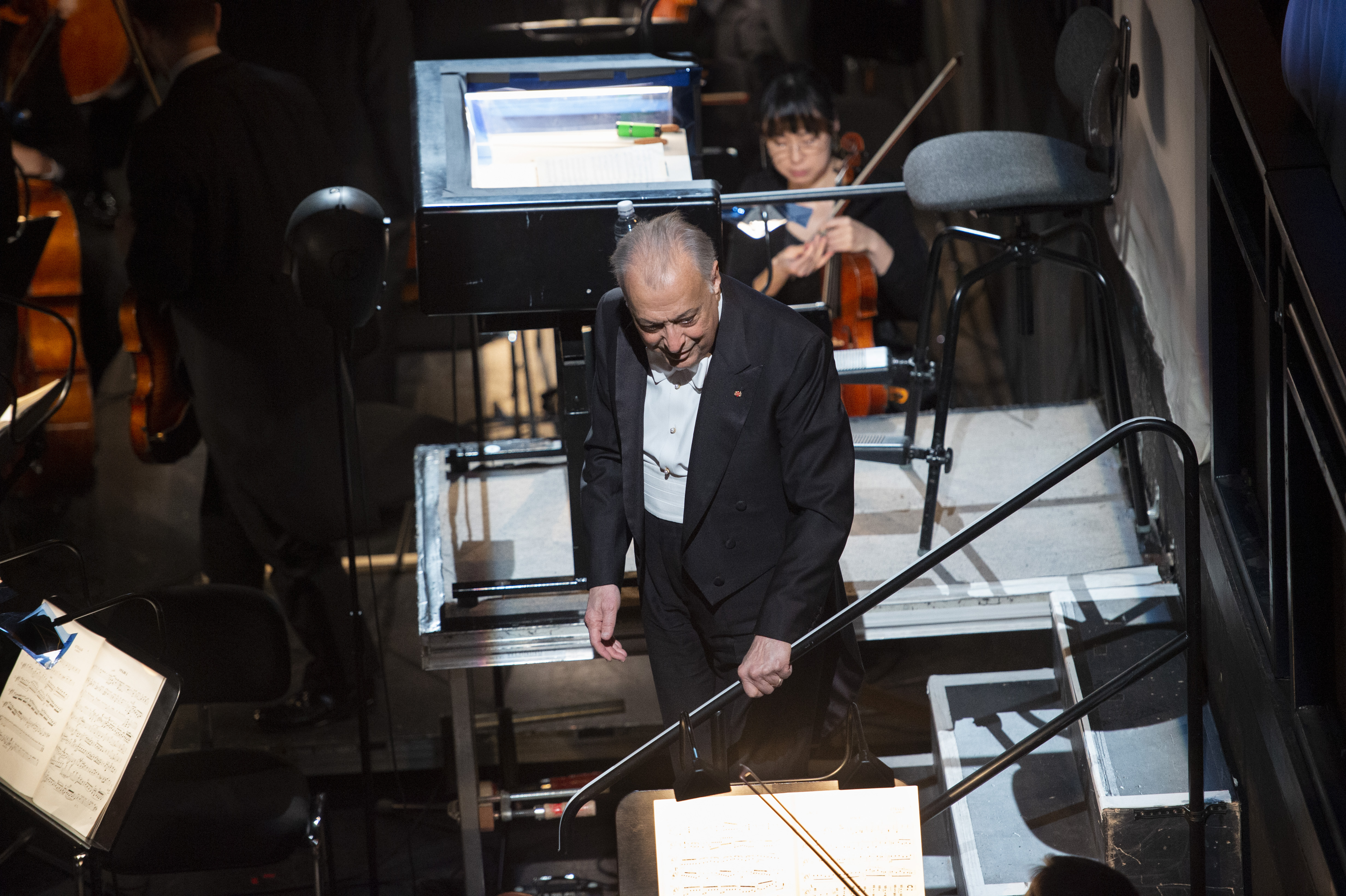Ein Dirigent im schwarzen Smoking steht auf einem Podium und blickt auf die Musiker in einem Orchestergraben. Ein Geiger sitzt hinter ihm, und Notenblätter sind auf Ständern zu sehen. Die Umgebung ist formell und schwach beleuchtet.