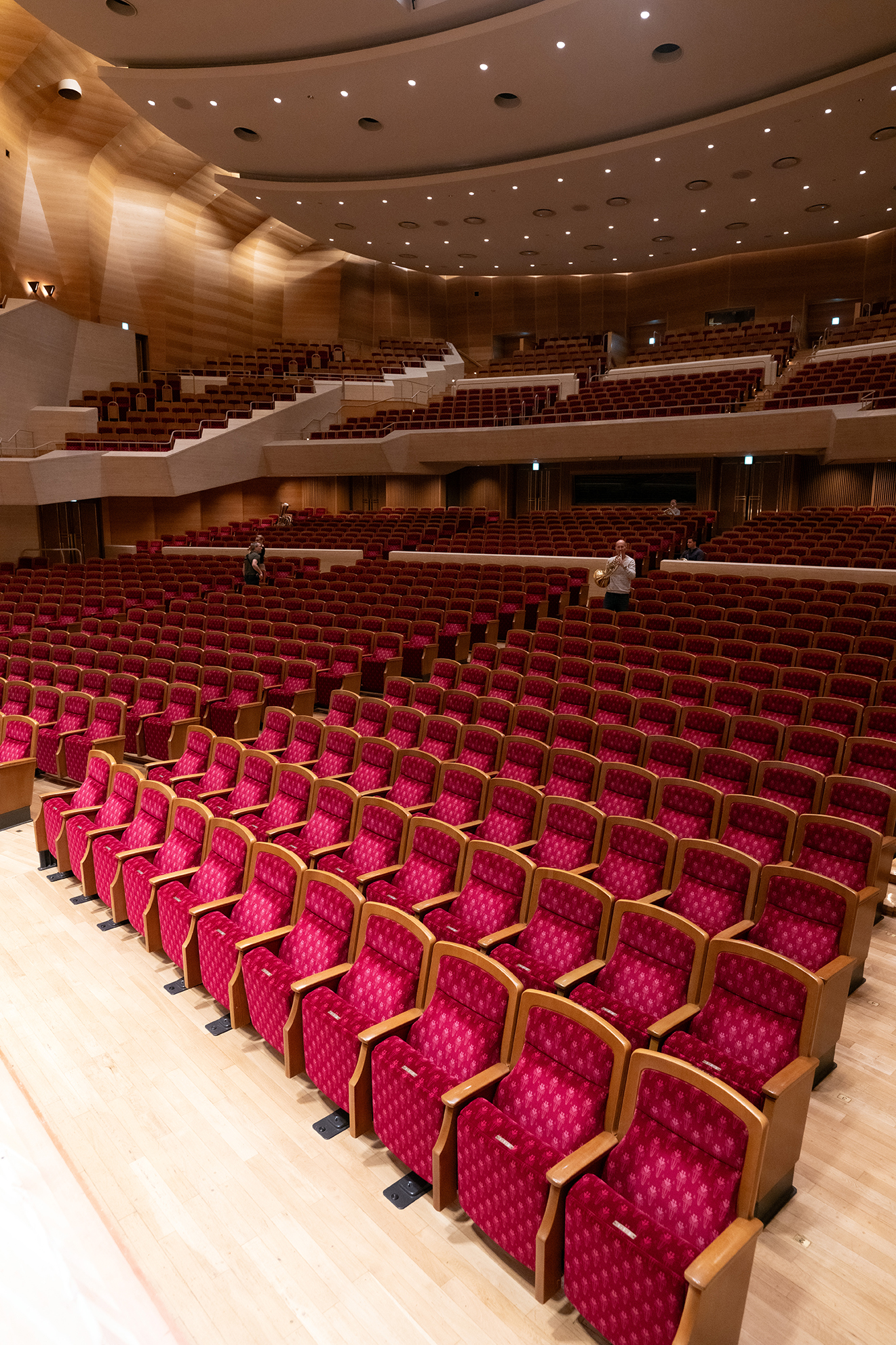 Ein großer, leerer Saal mit Reihen von rot gepolsterten Sitzen und hölzernen Armlehnen, einem hellen Holzboden und mehreren Balkonen. Ein paar Leute sind im Saal verstreut.